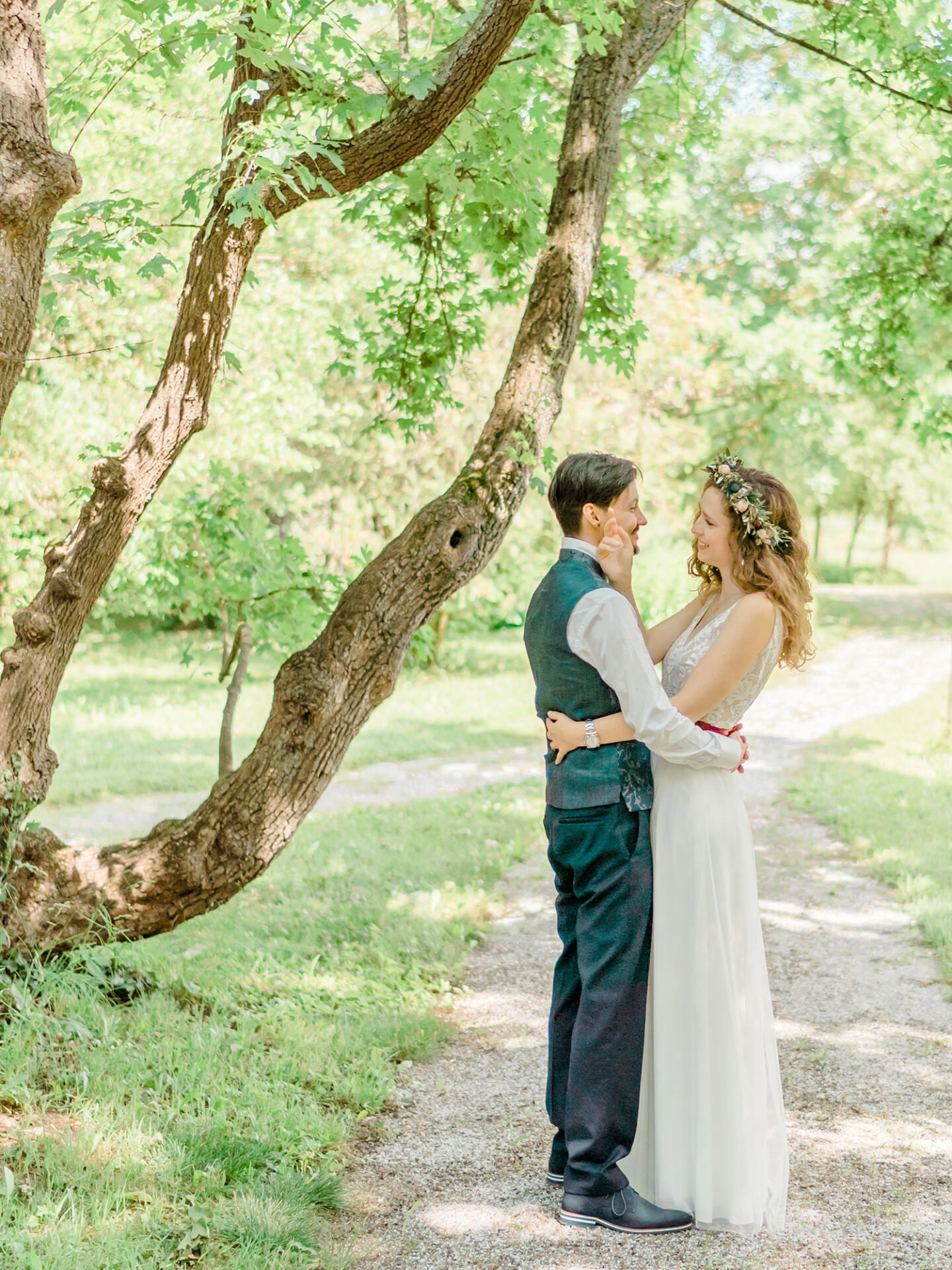 Braut und Bräutigam umarmen sich liebevoll auf einem ruhigen Waldweg. Die Braut mit einem Blumenkranz im Haar und der Bräutigam in einem eleganten Hochzeitsanzug stehen unter einem großen Baum, der die romantische Szene umrahmt.