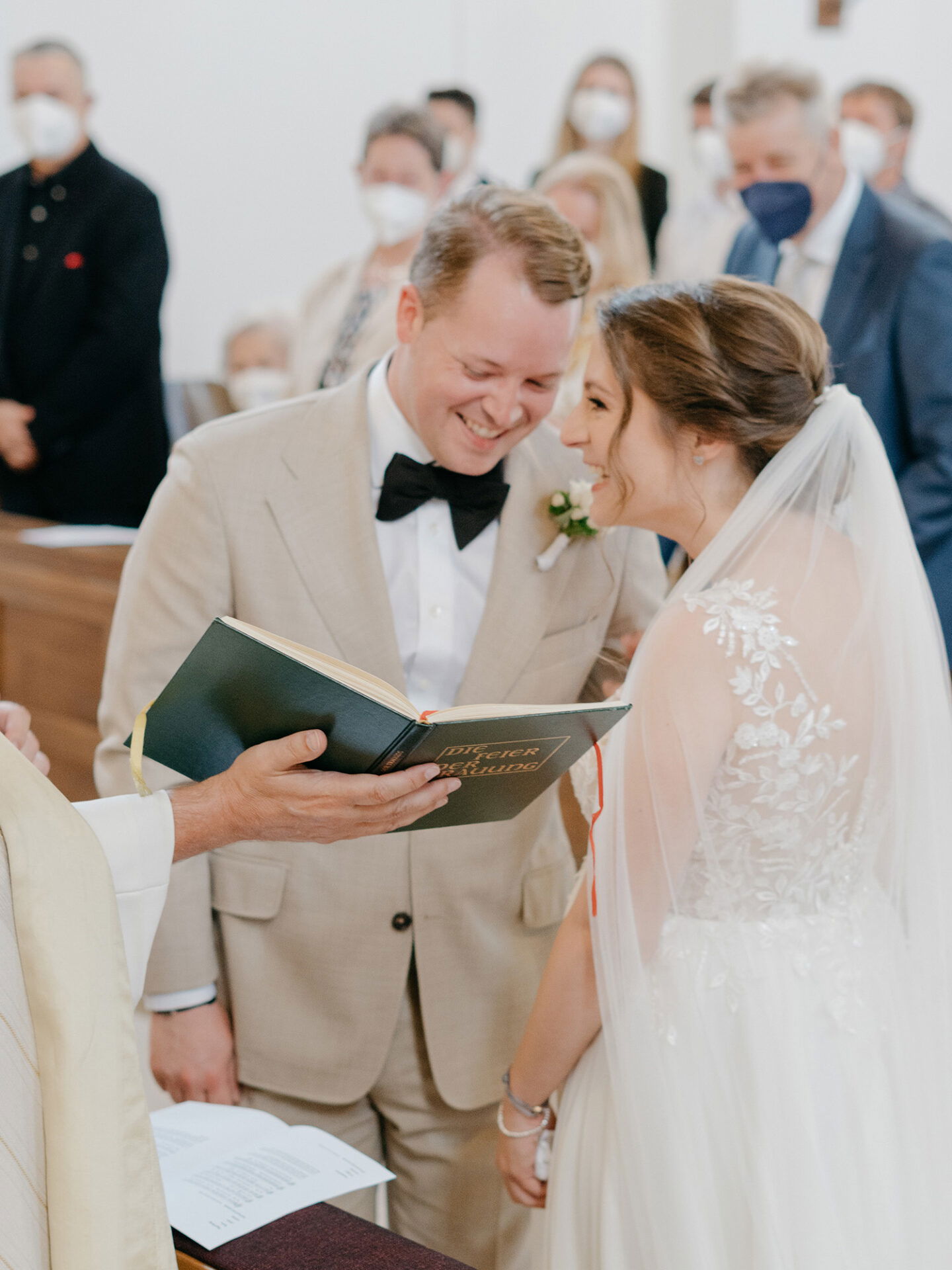 Ein emotionaler Moment während der kirchlichen Trauung: Das Brautpaar teilt ein Lächeln, während der Priester das Buch der Liturgie liest. Die Braut, in einem Spitzenkleid mit Schleier, und der Bräutigam in einem beigen Anzug sind von maskierten Gästen umgeben.