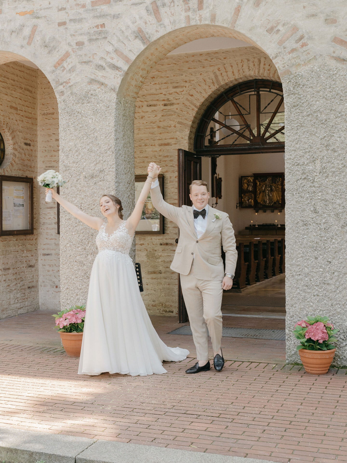 Das Brautpaar steht freudestrahlend vor einer rustikalen Kirche. Die Braut, in einem Spitzenhochzeitskleid, hebt triumphierend ihren Brautstrauß, während der Bräutigam in einem beigen Anzug sie stolz an der Hand hält.