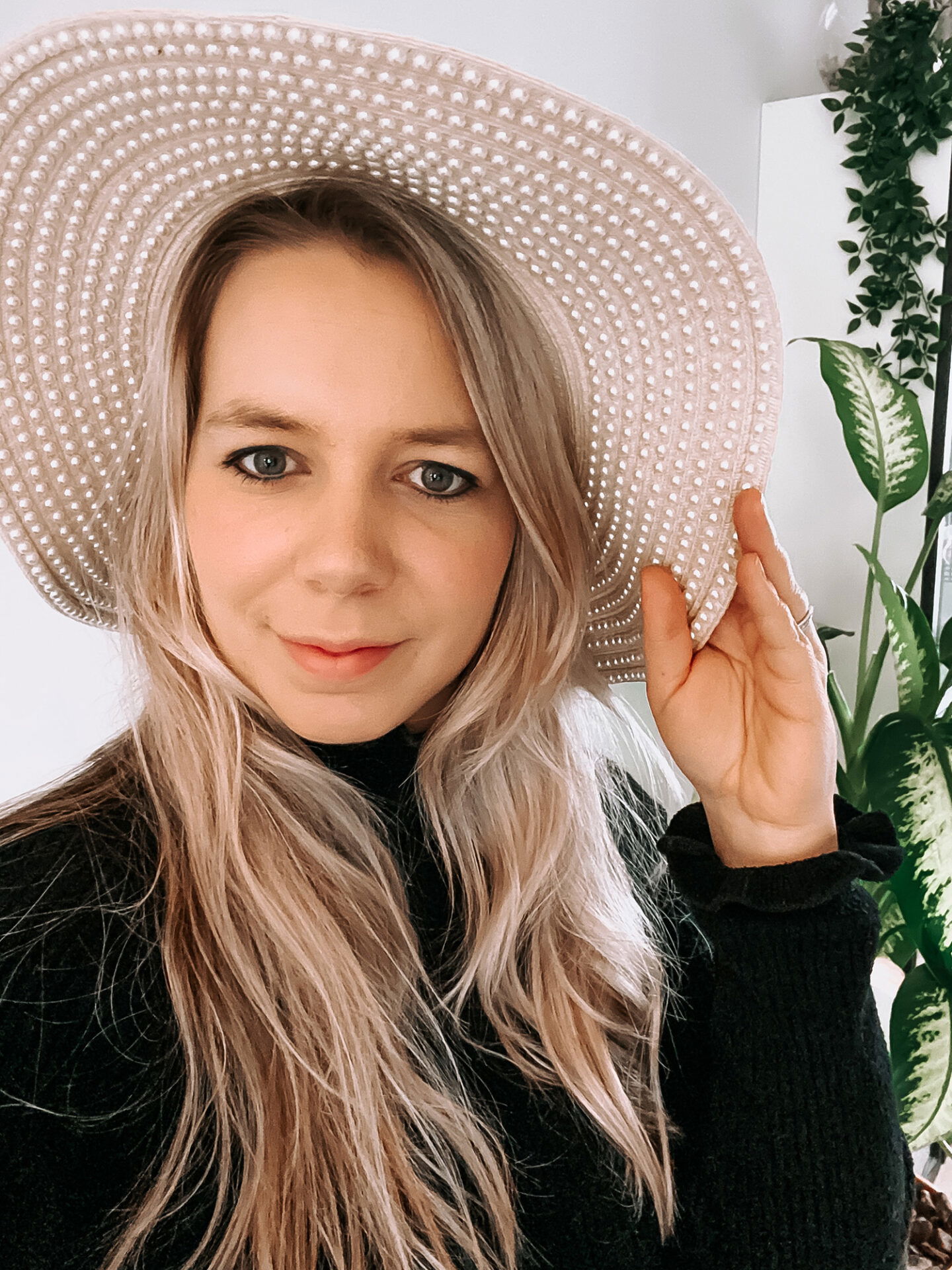 Eine Frau mit blonden Haaren und einem perlenverzierten Strohhut posiert vor einer Pflanze im Hintergrund. Sie hält den Rand des Hutes fest und zeigt ein dezentes Lächeln.