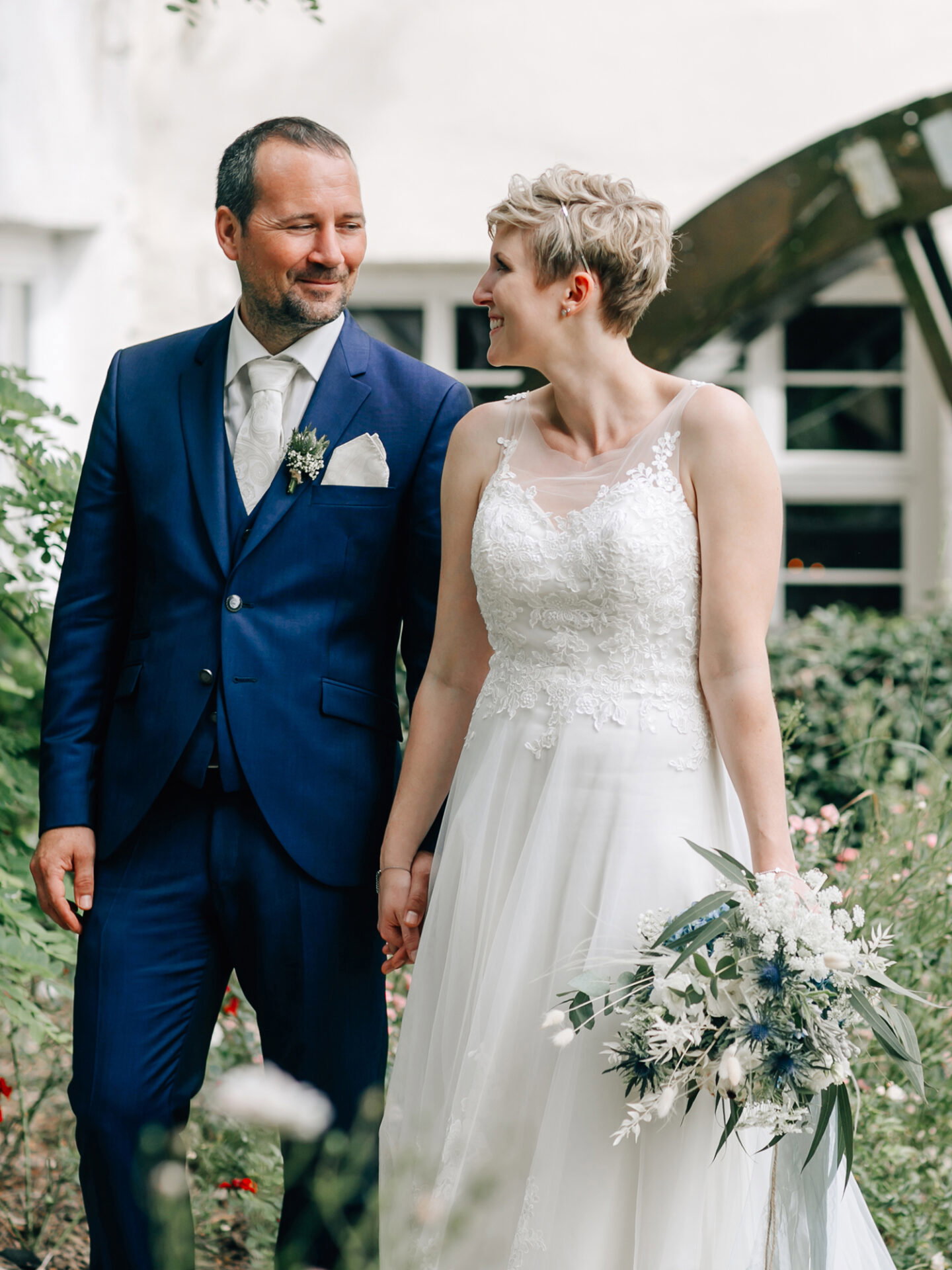 Ein Bräutigam im dunkelblauen Anzug und eine Braut im weißen Spitzenkleid gehen Hand in Hand durch einen mit Blumen bewachsenen Außenbereich. Die Braut trägt einen natürlichen Blumenstrauß mit grünen Blättern und weißen Blüten, während das Paar sich liebevoll anlächelt.
