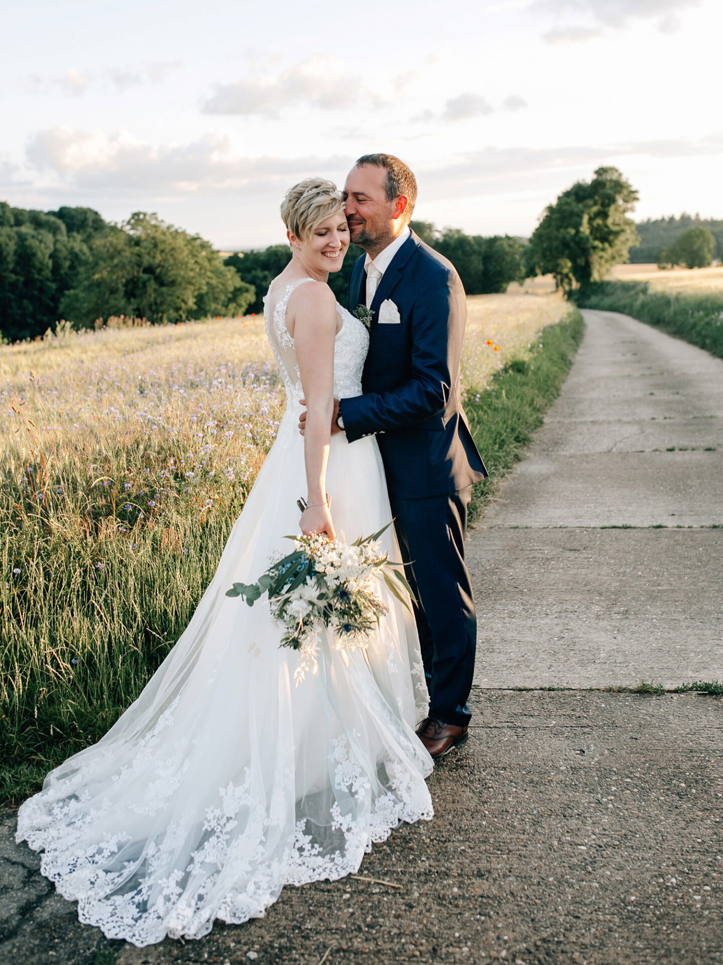 Braut und Bräutigam stehen auf einem Pfad neben einer wilden Wiese. Die Braut trägt ein langes, detailreiches Kleid mit Spitze und einen üppigen Blumenstrauß, während der Bräutigam in einem dunklen Anzug sie zärtlich von hinten umarmt. Die goldene Abendsonne taucht die Szene in ein warmes Licht.