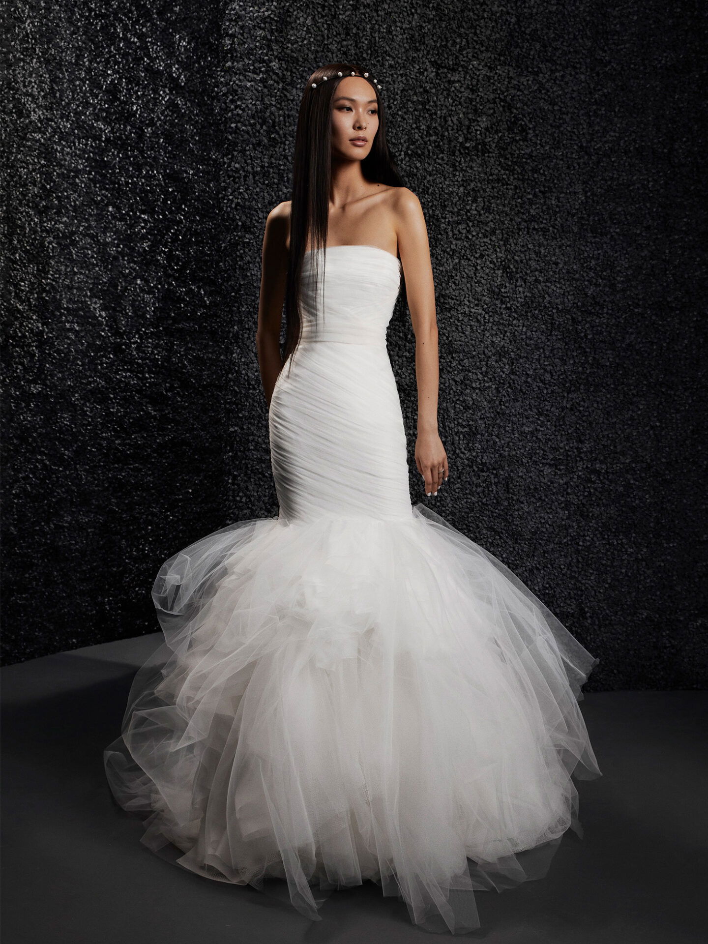 Ein modernes Brautkleid im Meerjungfrauenstil, das an der Taille eng anliegt und in einen ausladenden Tüllrock übergeht, verleiht der Braut eine dramatische Ausstrahlung. Der dunkle, strukturierte Hintergrund kontrastiert mit dem hellen, eleganten Kleid.