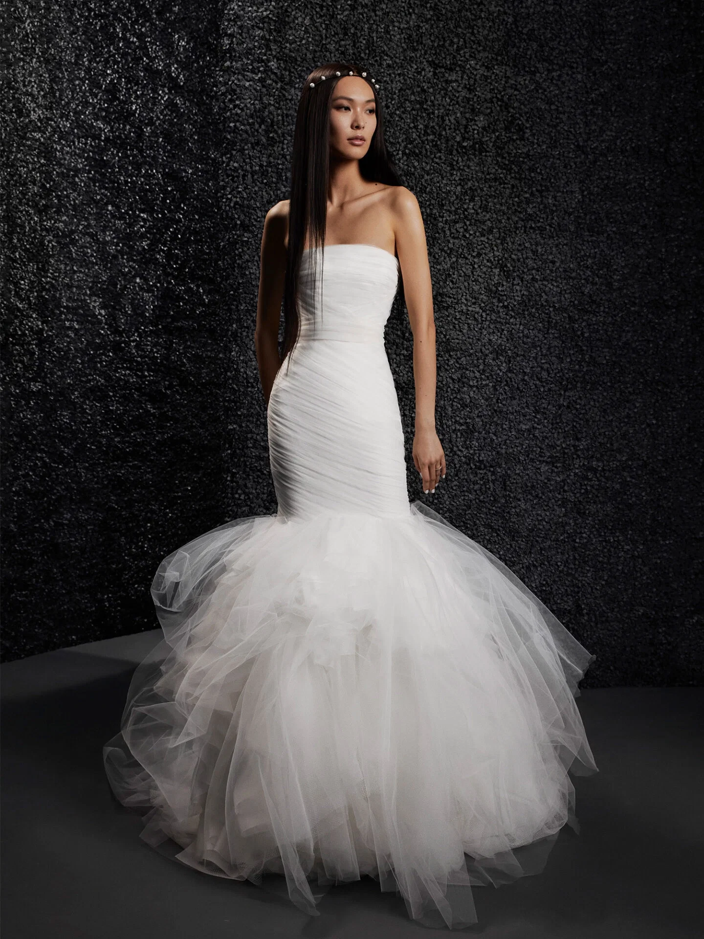 Ein modernes Brautkleid im Meerjungfrauenstil, das an der Taille eng anliegt und in einen ausladenden Tüllrock übergeht, verleiht der Braut eine dramatische Ausstrahlung. Der dunkle, strukturierte Hintergrund kontrastiert mit dem hellen, eleganten Kleid.