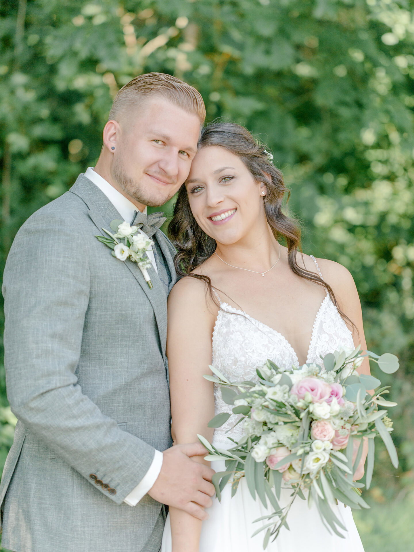 Das Brautpaar steht entspannt nebeneinander, der Bräutigam trägt einen grauen Anzug und die Braut ein weißes, spitzenbesetztes Kleid. In ihren Händen hält sie einen Blumenstrauß mit Eukalyptusblättern und zarten Blüten, umgeben von üppigem Grün im Hintergrund.