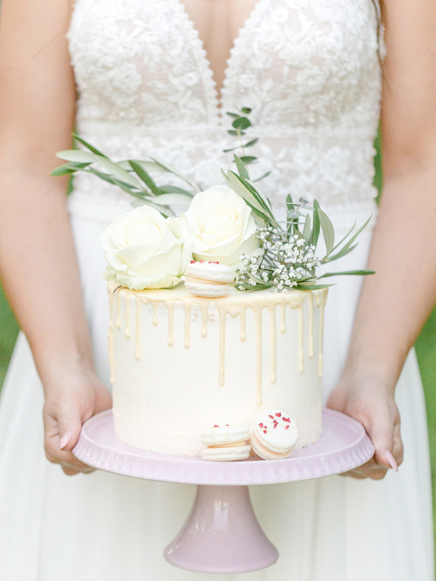 Eine elegante weiße Hochzeitstorte mit Macarons und weißen Rosen als Dekoration wird von einer Braut in einem Spitzenbrautkleid präsentiert. Die tropfende Glasur und die dezente Blumen- und Grünverzierung machen die Torte zum Highlight auf der rosa Tortenplatte.