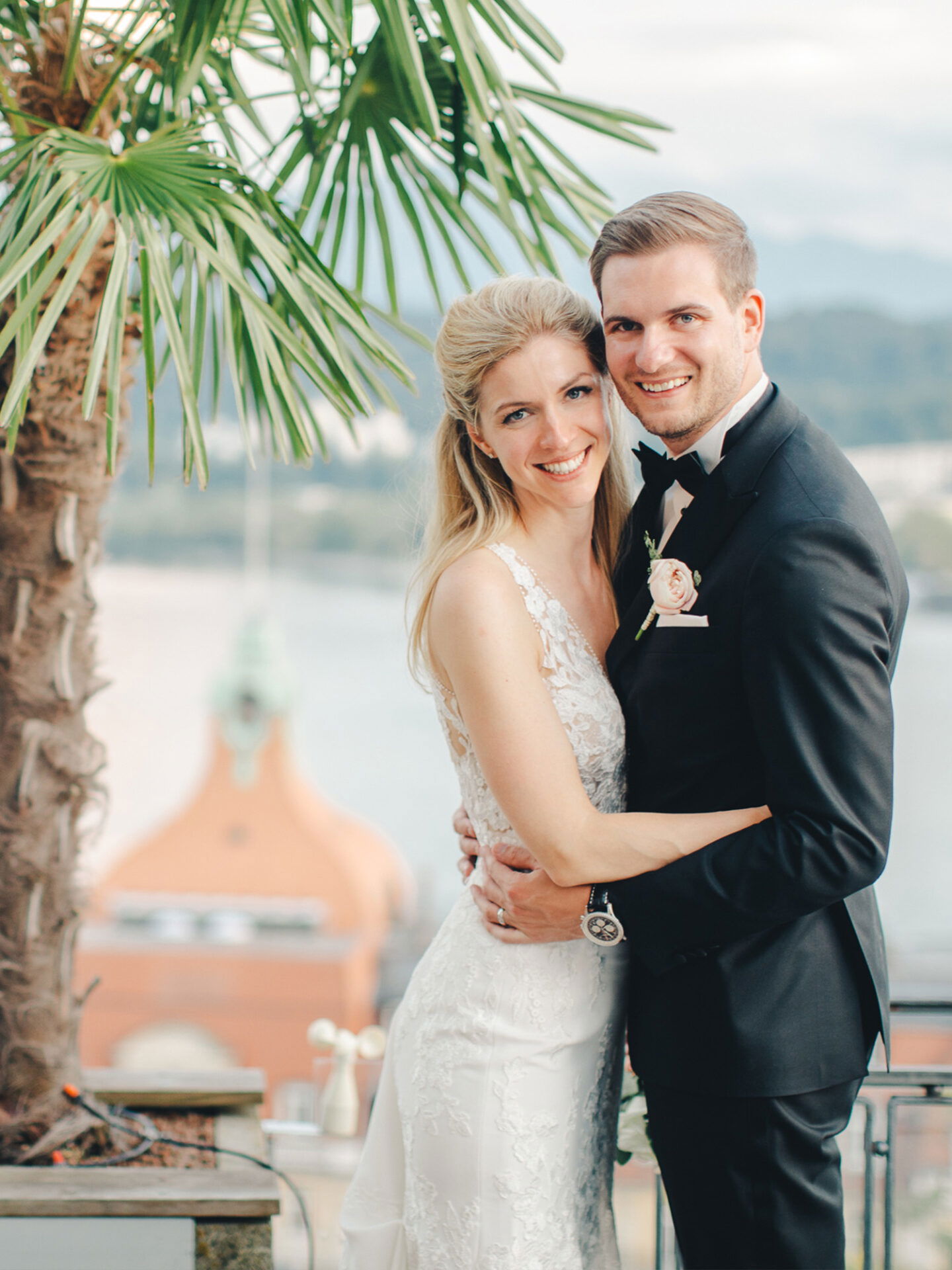 Ein strahlendes Brautpaar umarmt sich unter einer Palme, während sie in die Kamera lächeln. Die Braut trägt ein eng anliegendes Hochzeitskleid mit Spitze, der Bräutigam einen Smoking. Der Hintergrund zeigt einen Ausblick auf ein Ziegeldach und Berge.