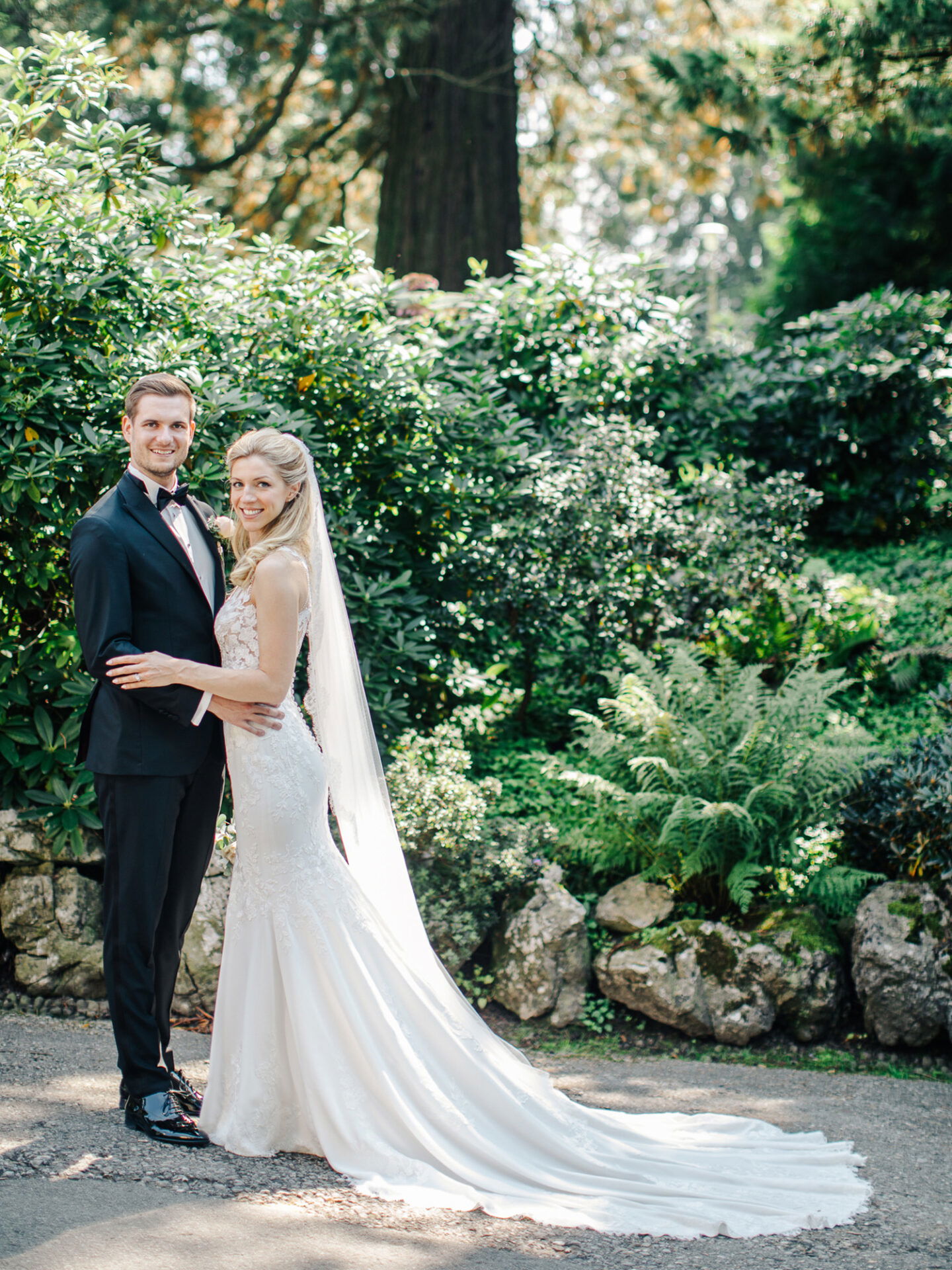 Das frisch verheiratete Paar posiert in einem grünen Garten mit dichter Vegetation. Die Braut trägt ein langes, fließendes Hochzeitskleid, während der Bräutigam in einem schwarzen Smoking und Fliege neben ihr steht. Beide wirken entspannt und glücklich.