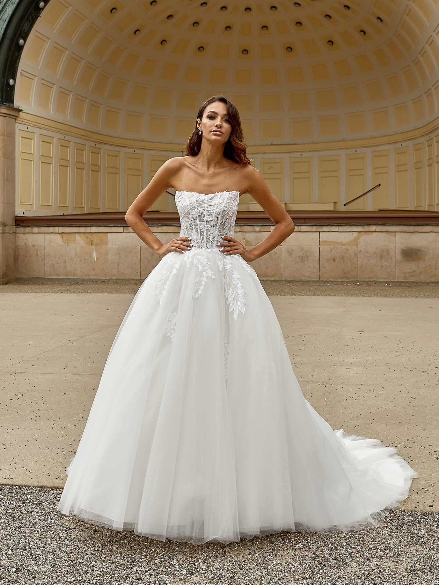 Das Bild zeigt eine Braut in einem modernen, trägerlosen Hochzeitskleid mit einem aufwendig verzierten Oberteil und fließendem Tüllrock. Im Hintergrund ist ein weitläufiger Pavillon mit gewölbtem Dach zu sehen, was dem Bild eine edle und klassische Atmosphäre verleiht.