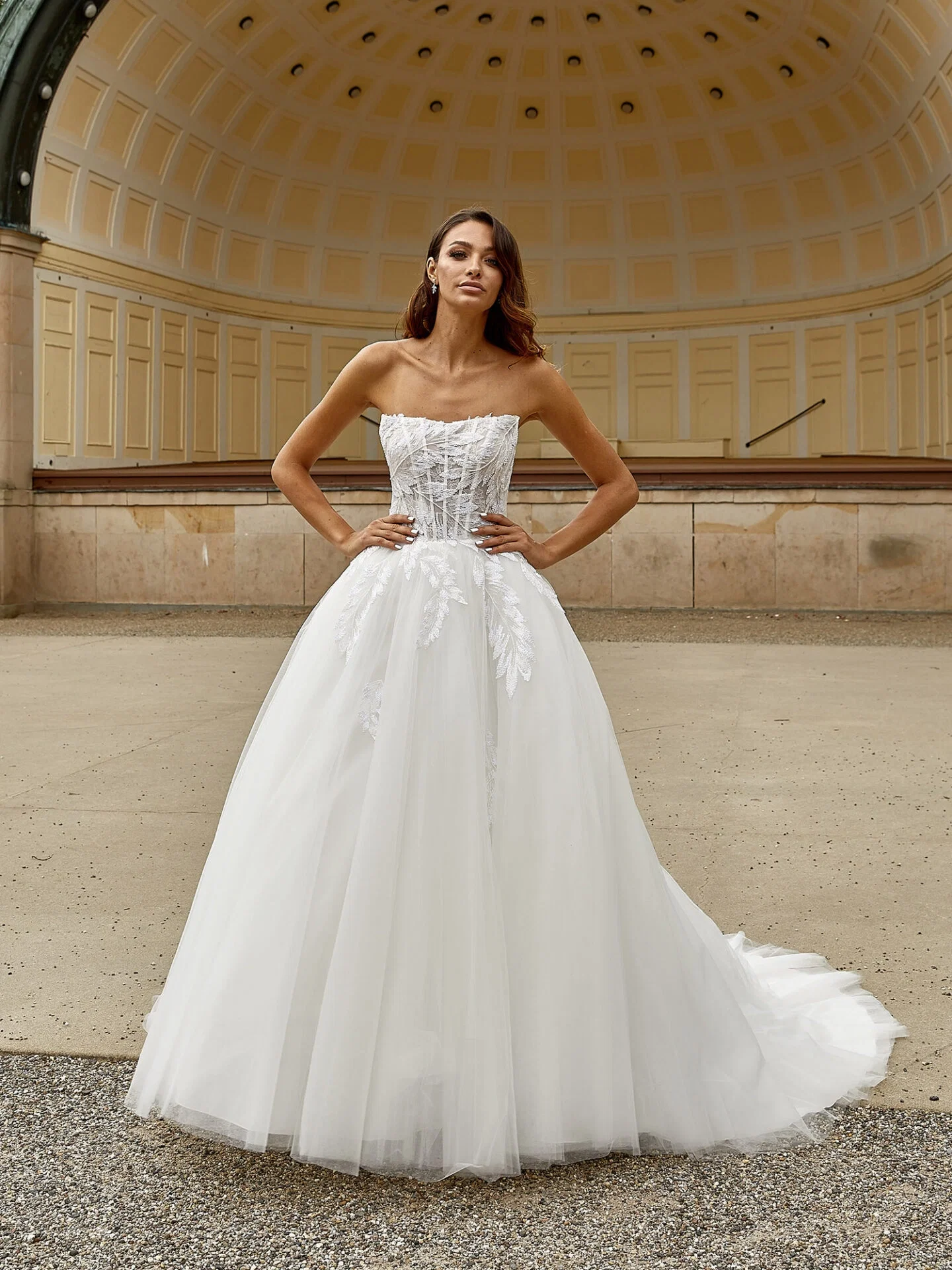 Das Bild zeigt eine Braut in einem modernen, trägerlosen Hochzeitskleid mit einem aufwendig verzierten Oberteil und fließendem Tüllrock. Im Hintergrund ist ein weitläufiger Pavillon mit gewölbtem Dach zu sehen, was dem Bild eine edle und klassische Atmosphäre verleiht.