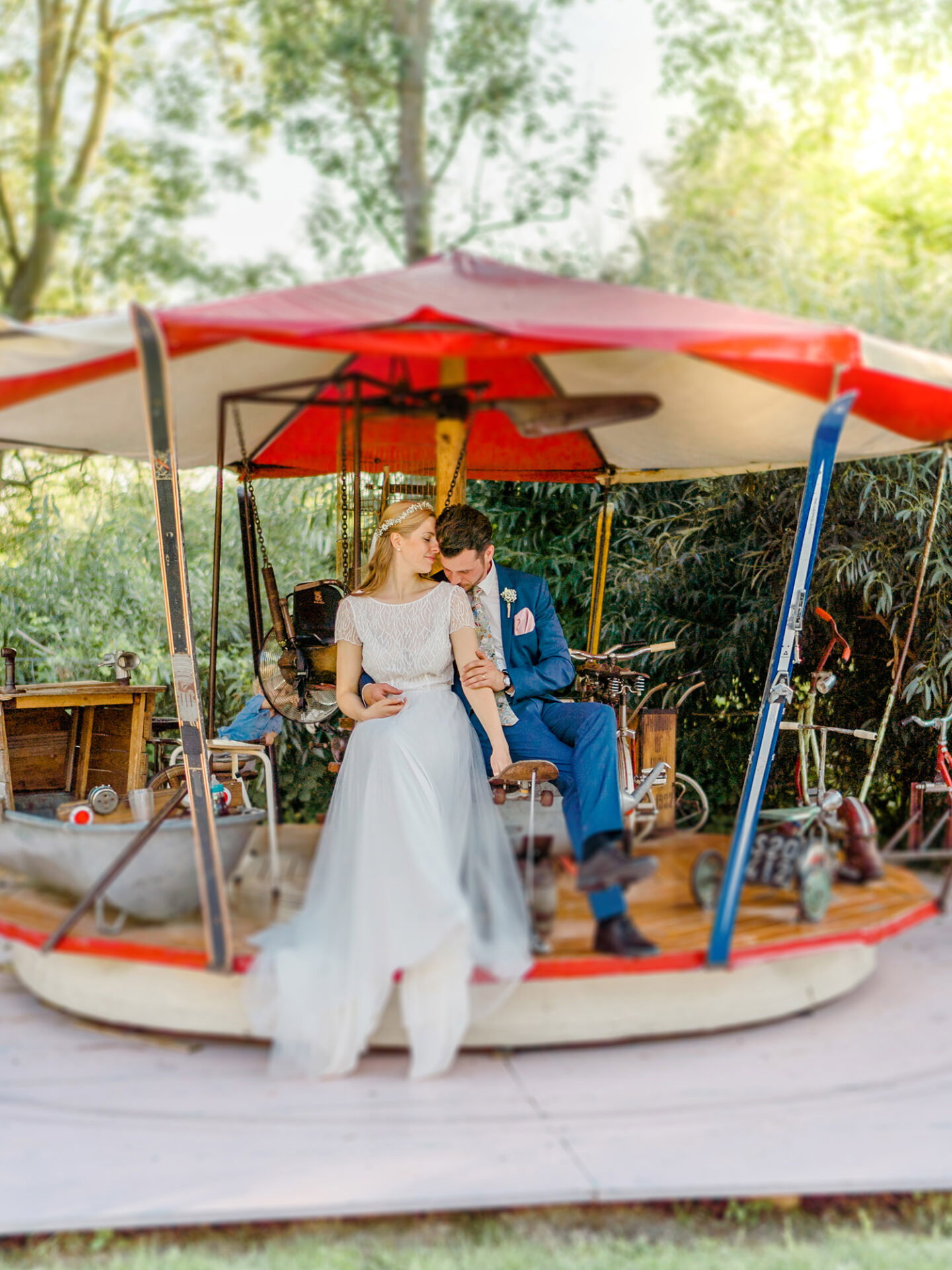 Braut und Bräutigam genießen einen ruhigen Moment auf einem historischen Karussell. Die Braut trägt ein schlichtes, aber wunderschönes Kleid, während der Bräutigam im blauen Anzug sie liebevoll an sich zieht. Das Karussell, umgeben von grüner Natur, wirkt wie ein Ort aus einer anderen Zeit.