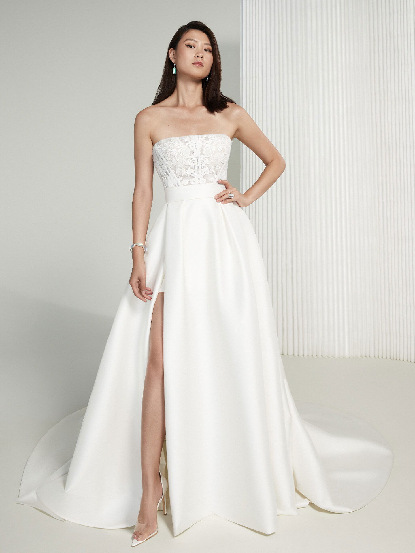 Eine Frau posiert selbstbewusst in einem stilvollen, trägerlosen Brautkleid mit floraler Spitze am Oberteil und einem tiefen Beinschlitz, der einen Hauch von Glamour und Modernität vermittelt.