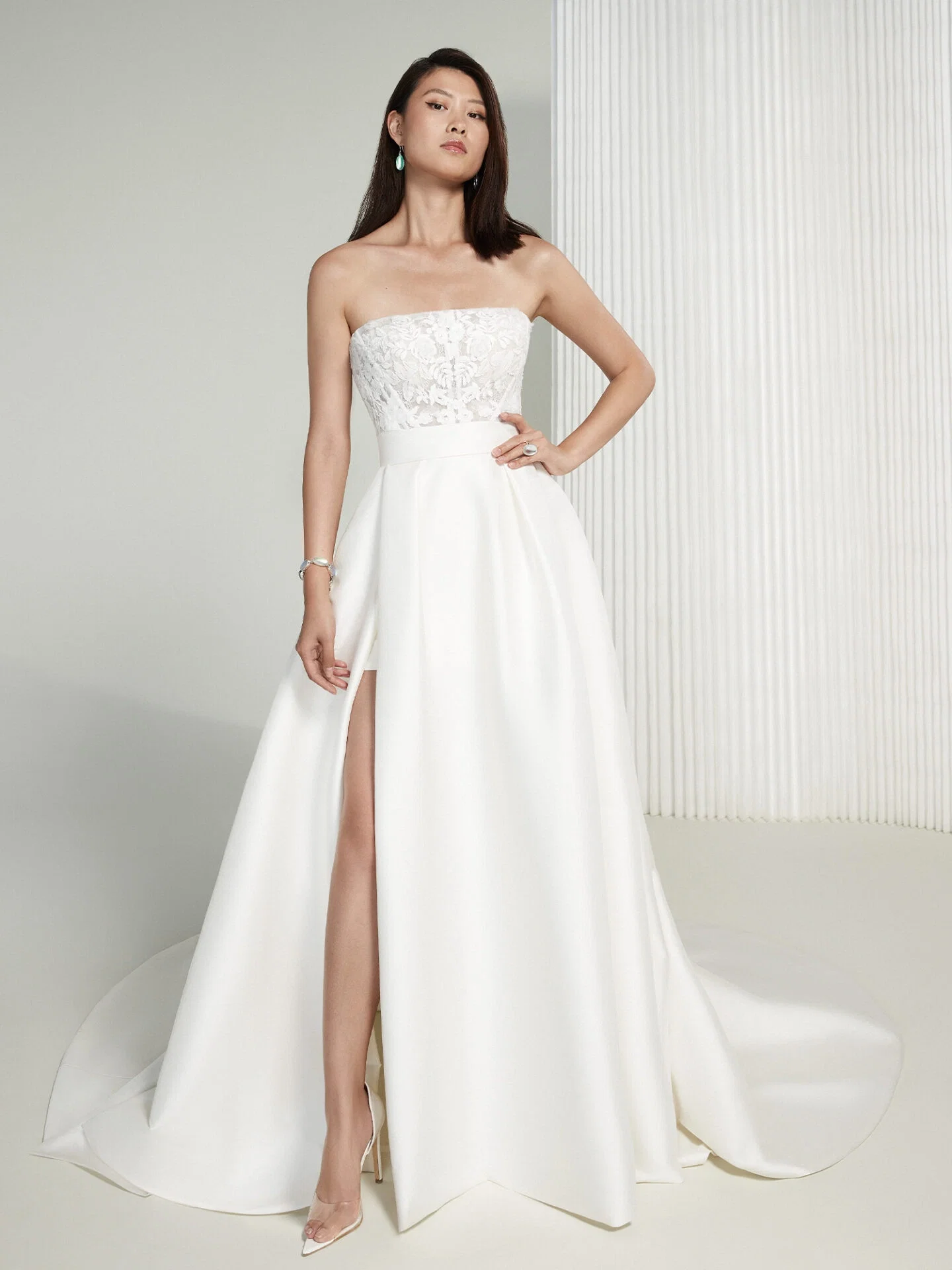 Eine Frau posiert selbstbewusst in einem stilvollen, trägerlosen Brautkleid mit floraler Spitze am Oberteil und einem tiefen Beinschlitz, der einen Hauch von Glamour und Modernität vermittelt.