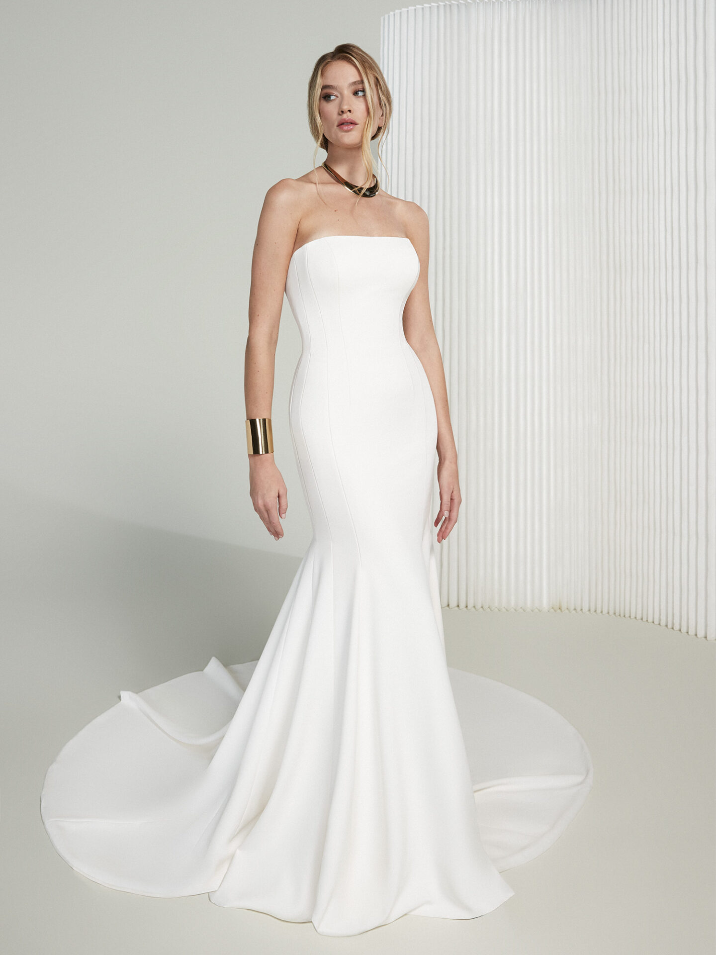 Ein trägerloses Hochzeitskleid im schlanken, minimalistischen Design mit einer dezenten Schleppe. Das schlichte Meerjungfrauen-Kleid schmeichelt der Figur der Braut. Goldene Schmuckstücke, wie eine breite Armspange und eine dezente Halskette, runden den eleganten Look ab.