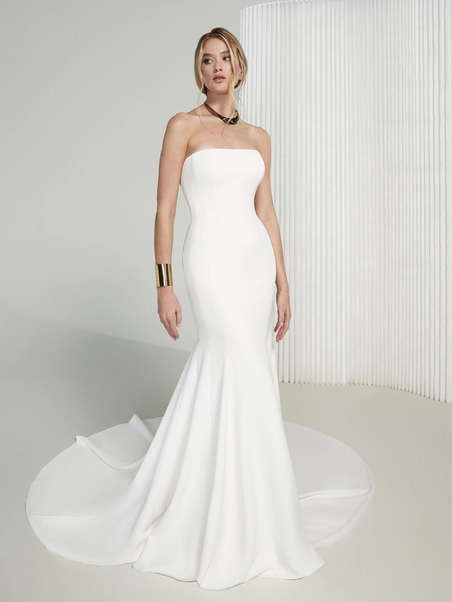 Ein trägerloses Hochzeitskleid im schlanken, minimalistischen Design mit einer dezenten Schleppe. Das schlichte Meerjungfrauen-Kleid schmeichelt der Figur der Braut. Goldene Schmuckstücke, wie eine breite Armspange und eine dezente Halskette, runden den eleganten Look ab.