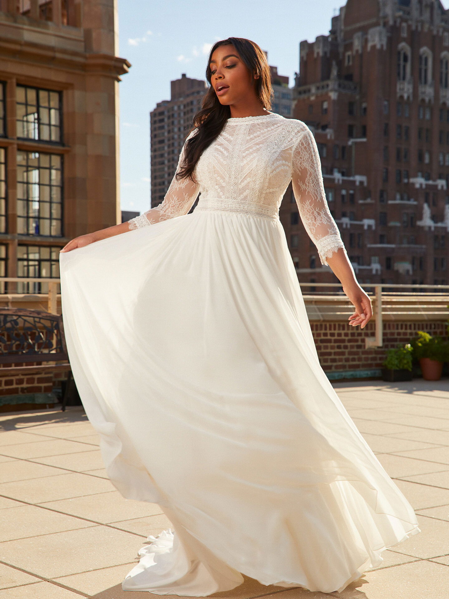 Eine Frau trägt ein Brautkleid mit langen Spitzenärmeln und einem fließenden, bodenlangen Rock, während sie auf einer städtischen Dachterrasse steht. Sie hebt den Saum ihres Kleides leicht an, wodurch eine zarte Bewegung des Stoffes entsteht. Hohe Gebäude rahmen die Szene im Hintergrund ein.