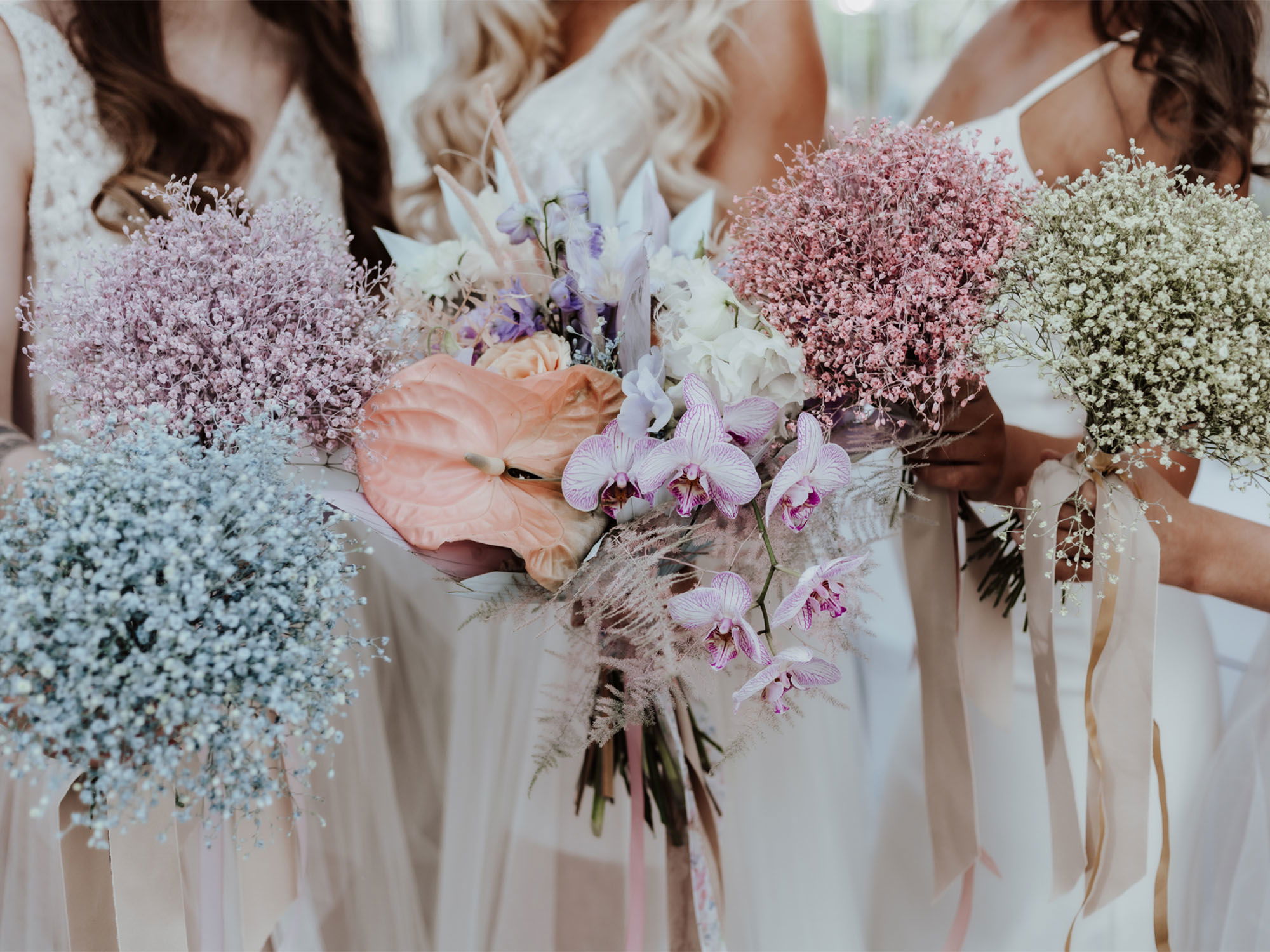 Bunte Brautsträuße mit zartem Schleierkraut und Orchideen in weichen Pastellfarben, die elegant von den Bräuten präsentiert werden.