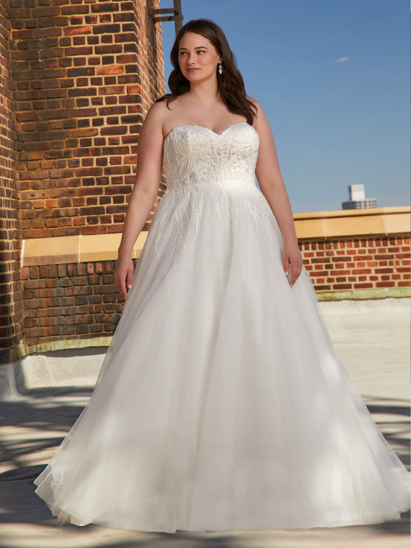 Eine Frau steht in einem eleganten, voluminösen Brautkleid mit herzförmigem Ausschnitt. Das Design des Kleides mit Stickereien auf dem Oberteil fügt sich harmonisch in die urbane Szenerie ein, die durch eine Backsteinfassade im Hintergrund geprägt ist.