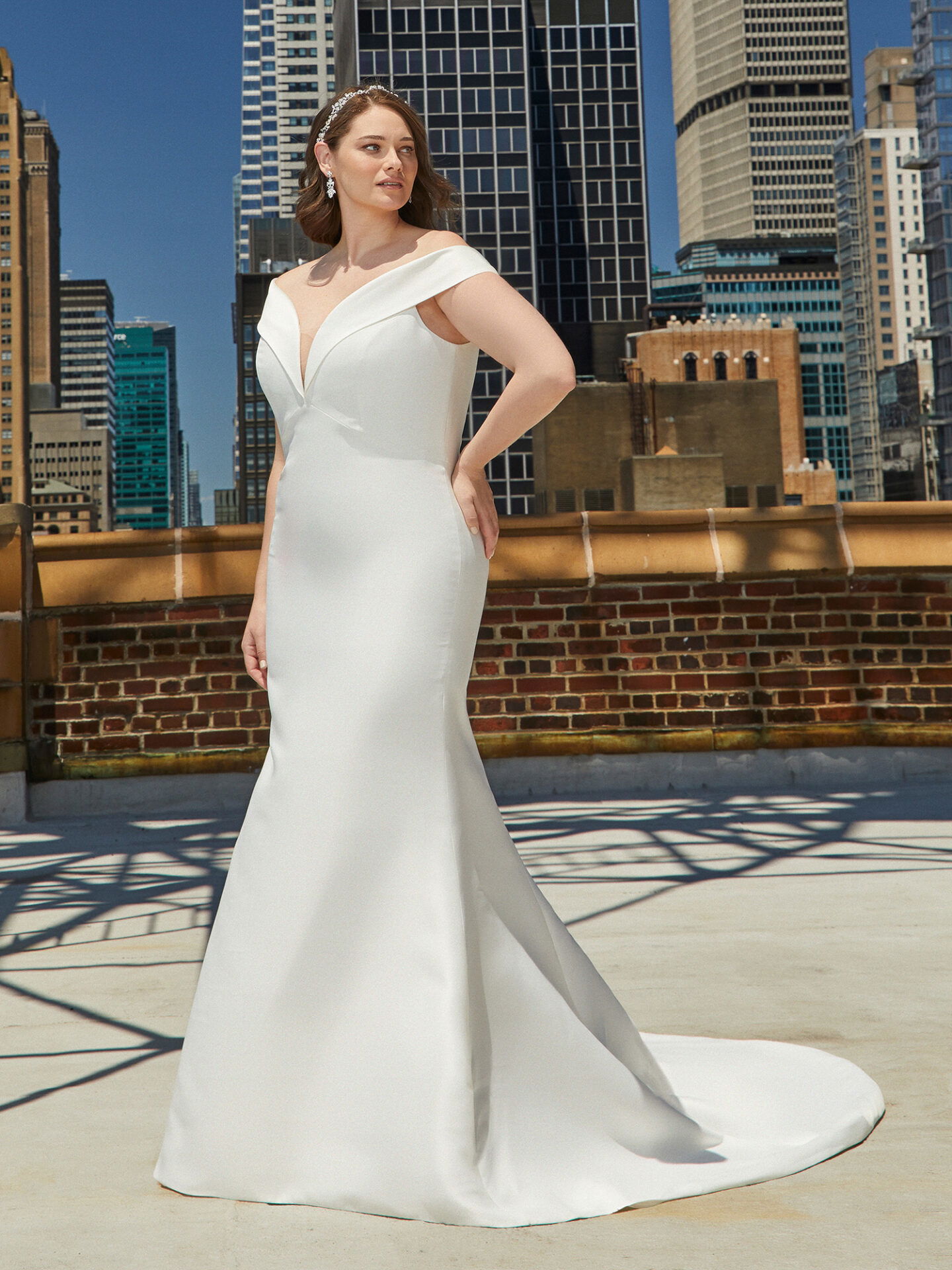 Eine Frau in einem Brautkleid mit figurbetontem Schnitt und einem breiten Ausschnitt steht stolz auf einer Dachterrasse. Der Hintergrund besteht aus modernen Bürogebäuden, die der Szene einen eleganten, städtischen Touch verleihen.