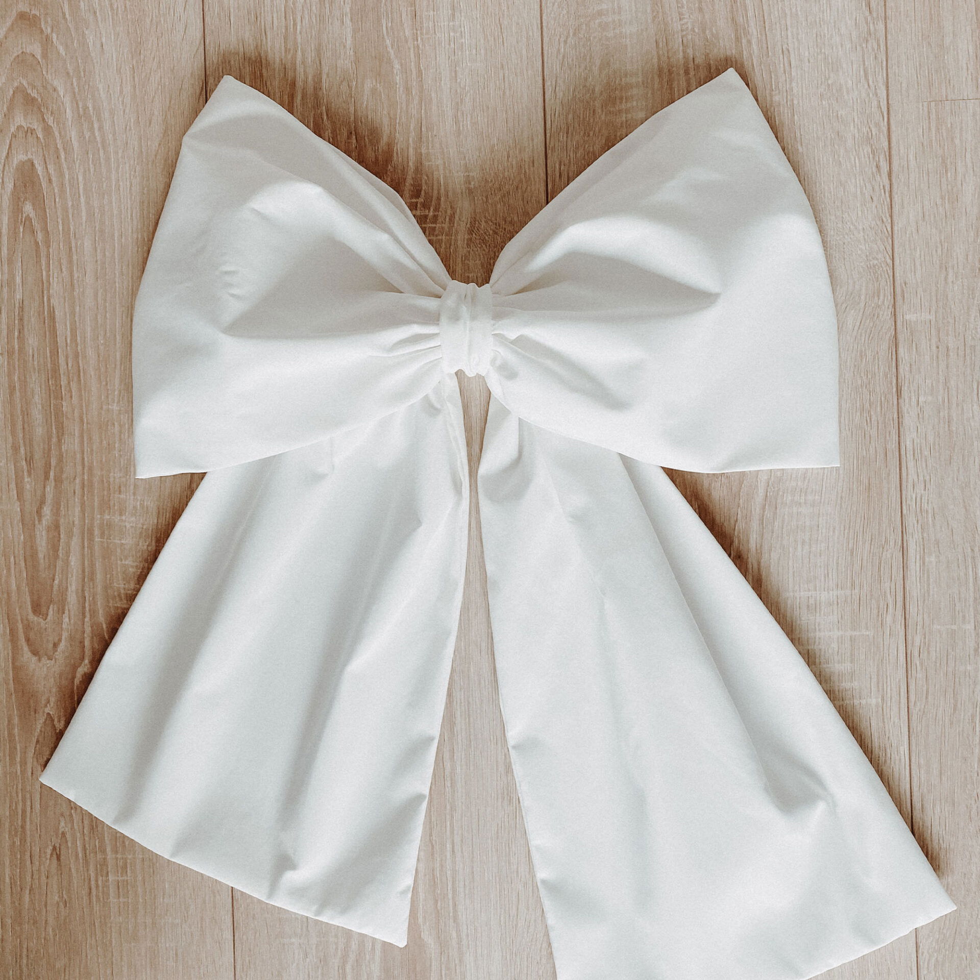 Eine flache, voluminöse weiße Schleife mit langen Bändern liegt auf einem hellen Holzboden. Die Schleife ist sauber gefaltet und glatt.