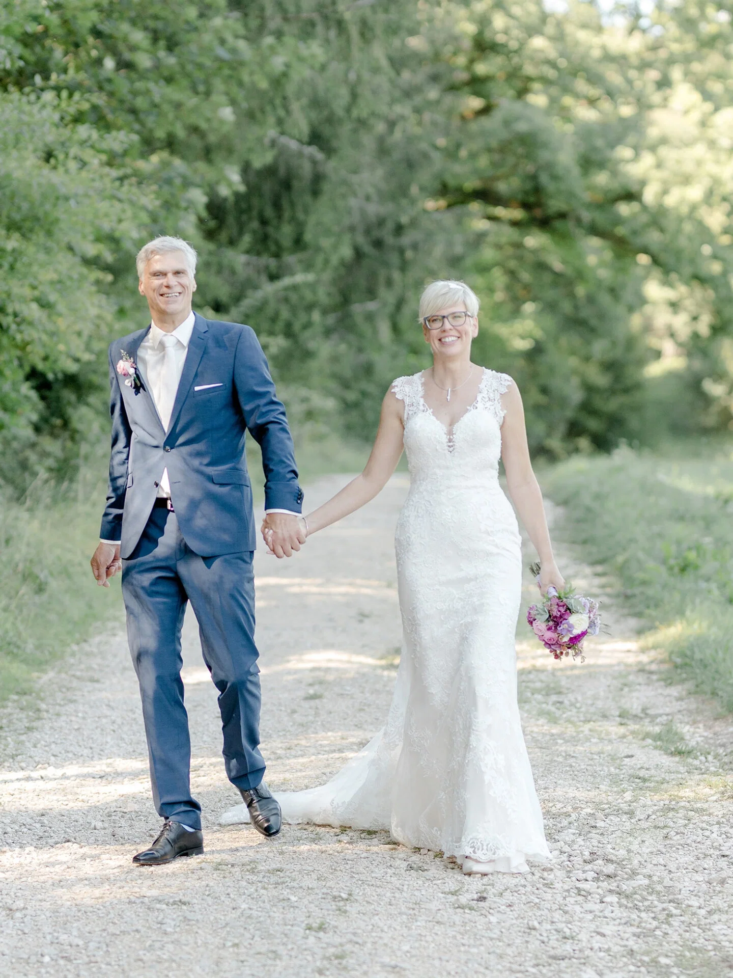 Sarah und Achim schreiten fröhlich einen mit Kies bedeckten Weg entlang, umgeben von üppigem Grün. Die Braut trägt ein bodenlanges, mit Spitzen versehenes Kleid und einen pink-violetten Blumenstrauß, während der Bräutigam einen klassischen blauen Anzug mit weißer Krawatte und einem Ansteckblumenstrauß trägt. Ihre entspannte Haltung und die ruhige Umgebung machen die Szene besonders romantisch.
