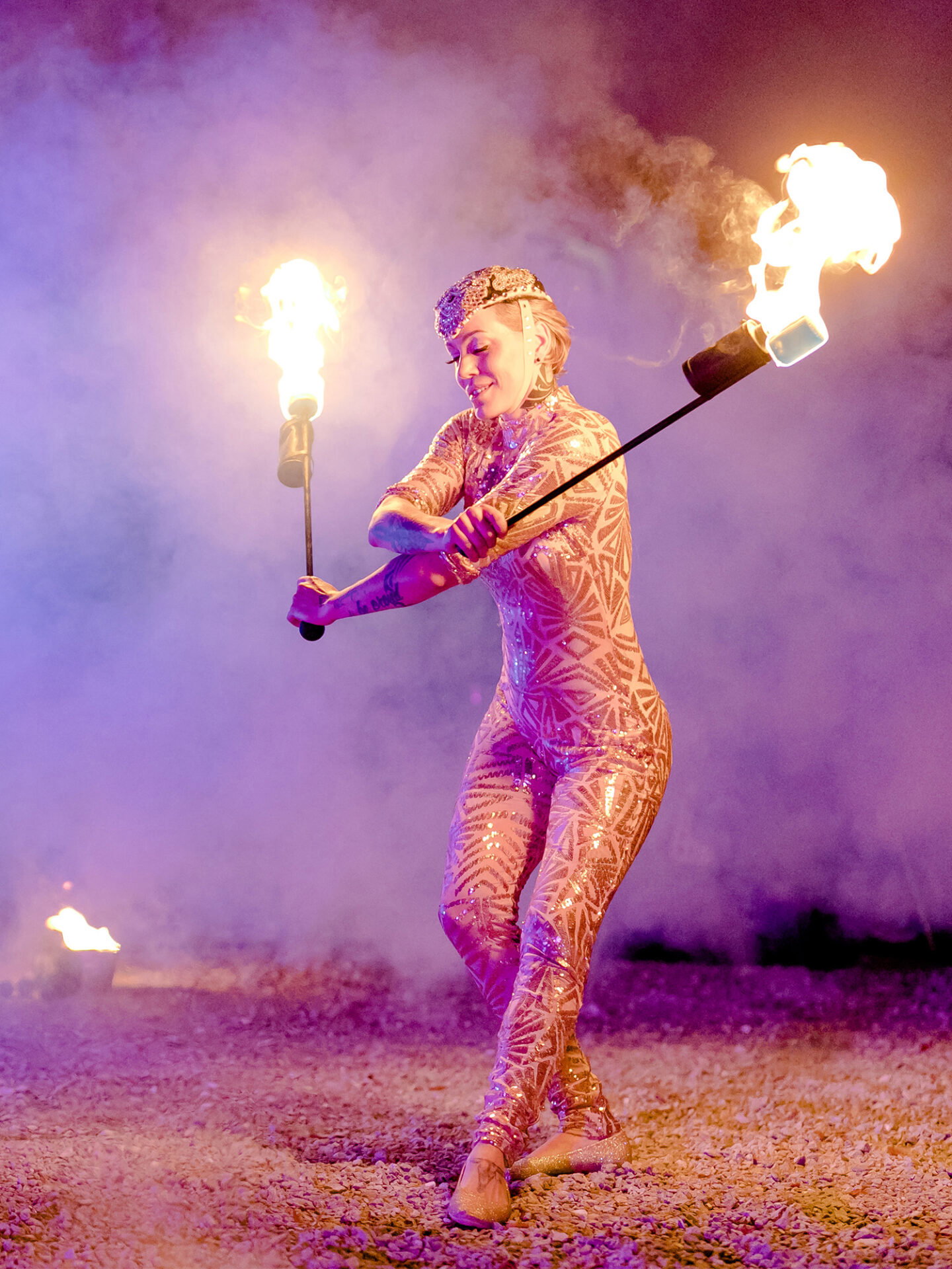 Eine Feuerartistin präsentiert ihre spektakuläre Darbietung mit brennenden Fackeln, während sie einen funkelnden Anzug mit komplizierten Mustern trägt. Die heiße Glut und der Rauch im Hintergrund verleihen der Szenerie einen magischen und intensiven Effekt, der das Publikum in den Bann zieht.