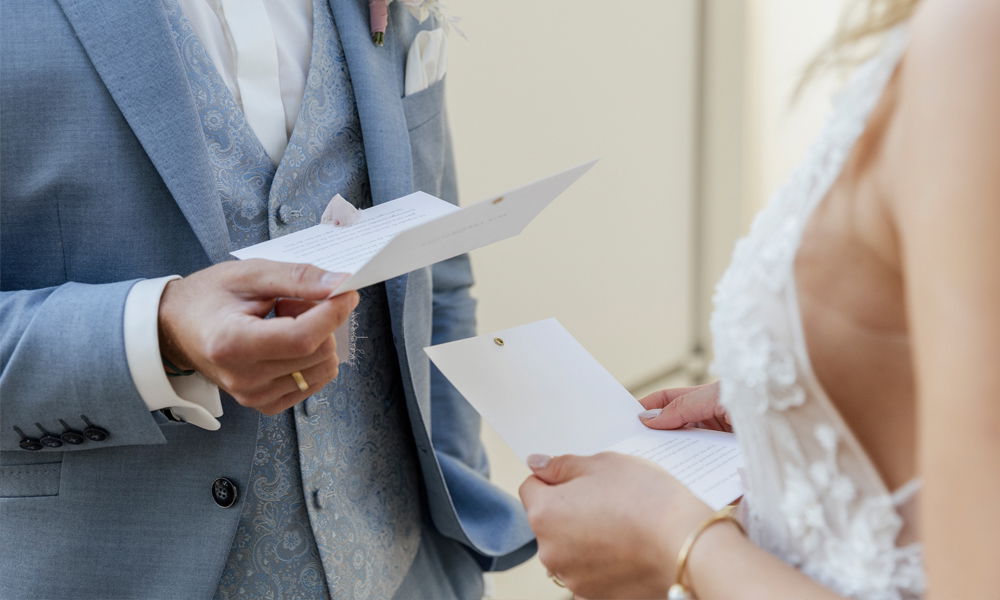 Zärtlicher Moment während der Trauung – das Brautpaar liest seine Eheversprechen, Fokus auf die Hände und feine Details der Kleidung.