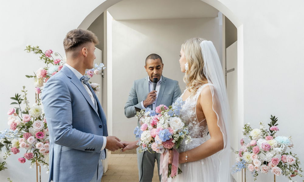 Trauung unter freiem Himmel mit einem Paar in hellblauem Anzug und floraler Spitze, der Redner spricht zwischen blühenden Gestecken.