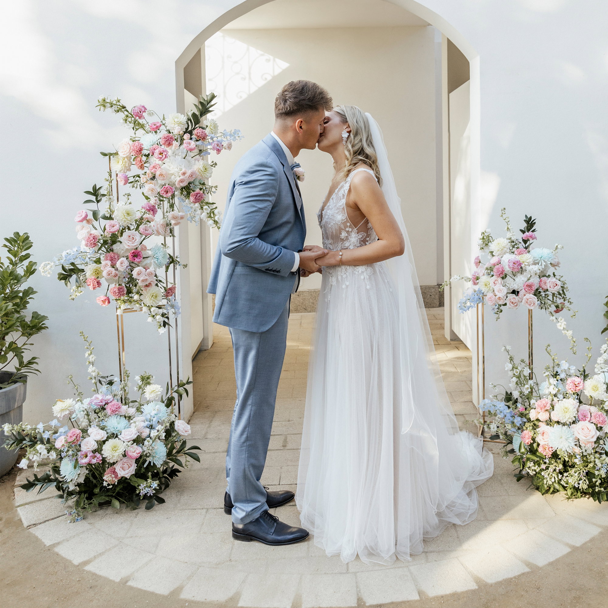 Hochzeitspaar beim Kuss im eleganten Außenbereich, der Bräutigam in hellblauem Anzug, die Braut in romantischem Spitzenkleid, eingerahmt von floralen Ständern in zarten Farben.