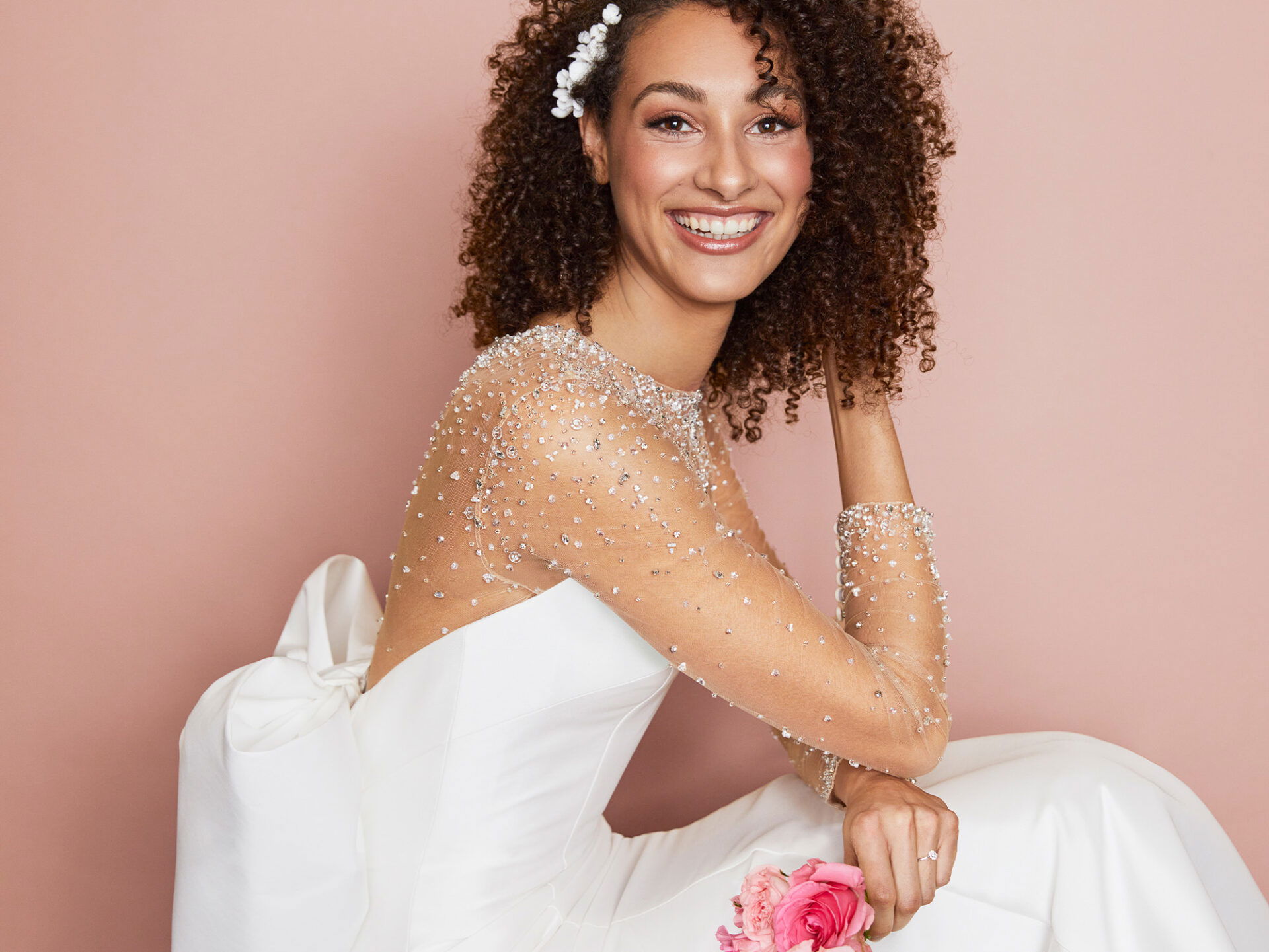 Eine Braut mit lockigem Haar trägt ein schulterfreies, weißes Hochzeitskleid mit funkelnden Details an den Ärmeln. Vor einem rosa Hintergrund posiert sie lächelnd, eine pinke Blume locker in ihrer Hand haltend.