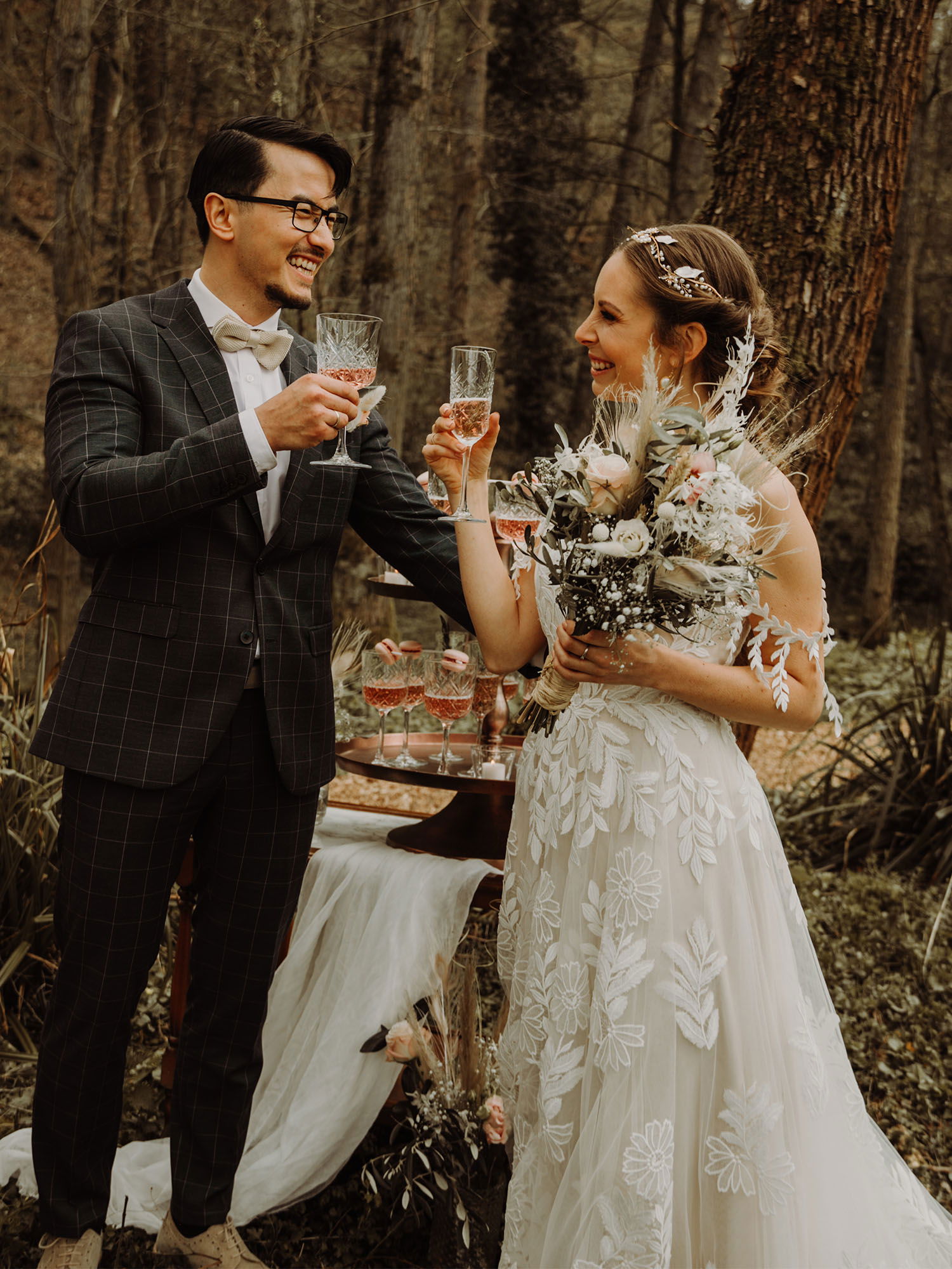 Fröhlicher Hochzeitstoast im Wald: Braut und Bräutigam lächeln sich an, während sie Sektgläser in die Luft halten. Die Braut trägt ein aufwendig besticktes Kleid und hält einen Blumenstrauß, während hinter ihnen ein eleganter Champagner-Turm steht.