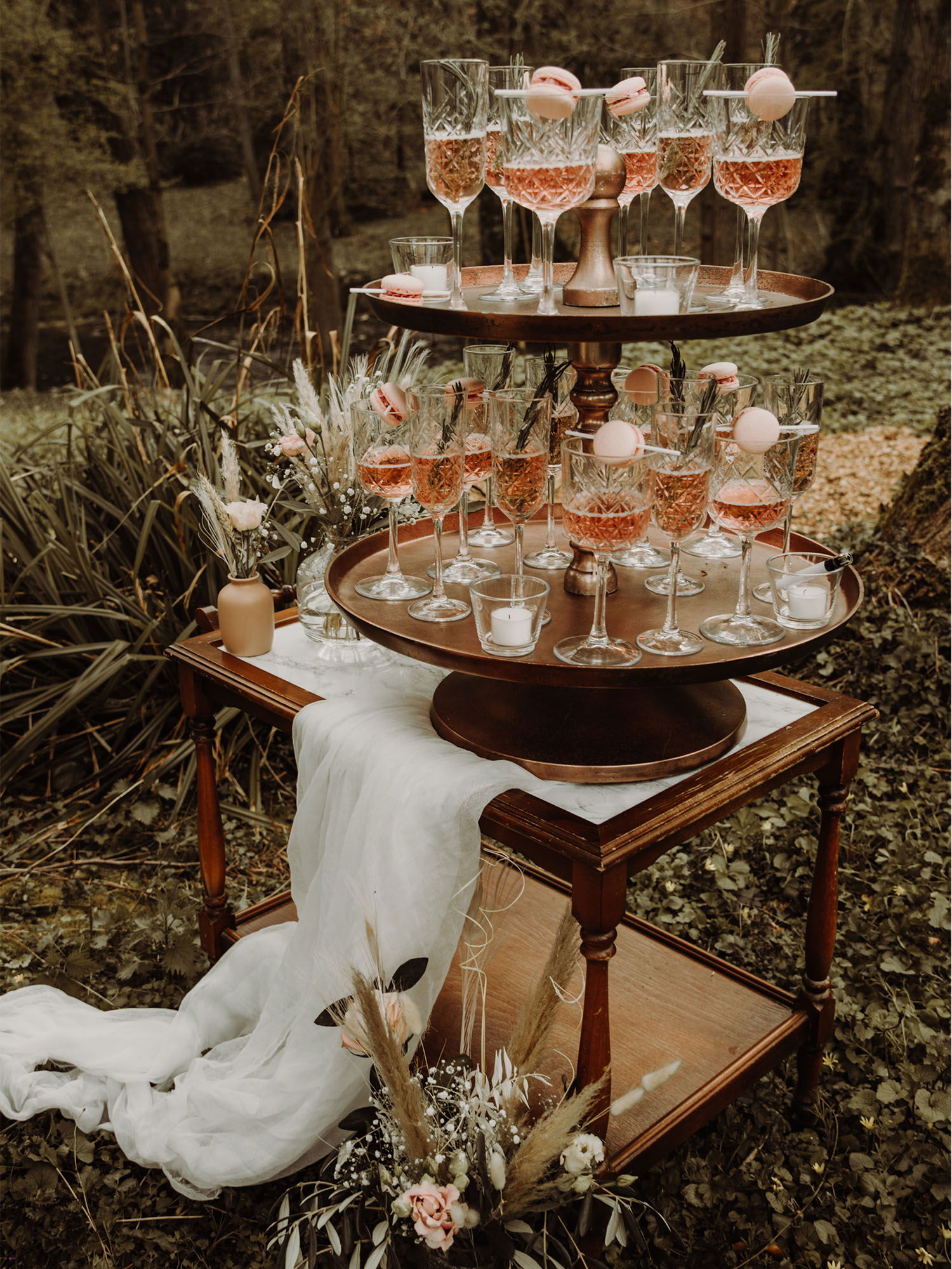 Zwei Ebenen eines Champagner-Turms im Wald, geschmückt mit Kristallgläsern voller Rosé-Sekt und rosa Macarons. Der Holztisch ist mit einem weißen Tuch drapiert, und die Umgebung aus grüner Vegetation sowie zarte Blumenarrangements vervollständigen die romantische Atmosphäre.