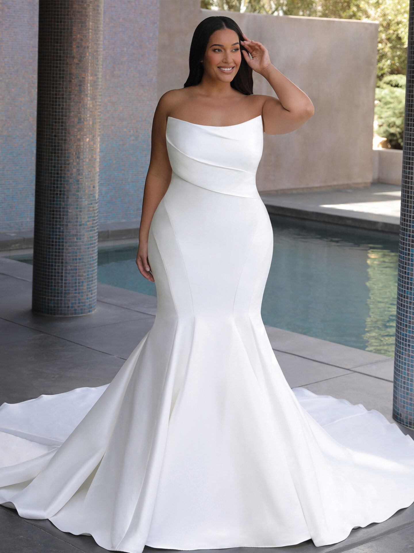 Eine strahlende Braut in einem klassischen, schulterfreien Hochzeitskleid posiert vor einem Pool. Das schlichte Design des Kleides mit seinem fließenden Rock und der klaren Linienführung hebt ihre Silhouette hervor.