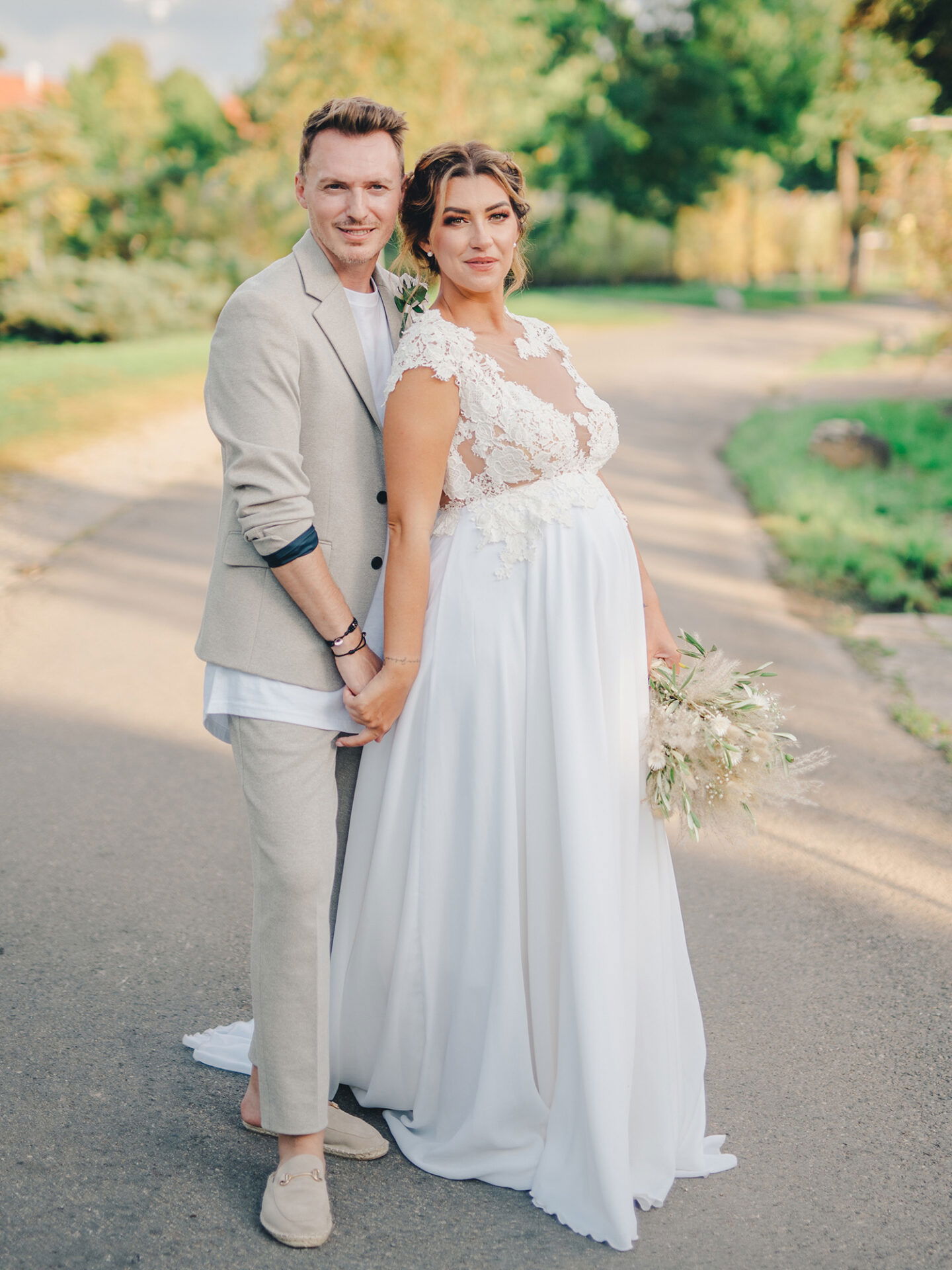 Die Braut, in einem weißen Spitzenhochzeitskleid, und der Bräutigam, in einem entspannten, hellen Anzug, stehen Hand in Hand auf einem ruhigen Weg. Sie lächeln in die Kamera, umgeben von der Natur an einem sonnigen Tag.