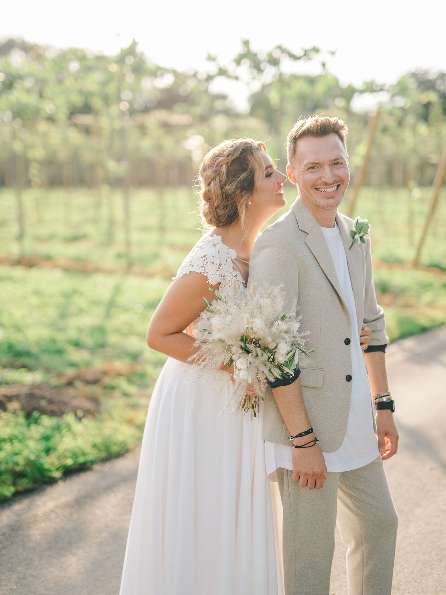 Die Braut und der Bräutigam genießen einen fröhlichen Moment im Freien. Die Braut trägt ein weißes Kleid mit Blumenstrauß, und der Bräutigam ist leger in einem hellen Anzug gekleidet. Sie stehen auf einem sonnenbeschienenen Weg.