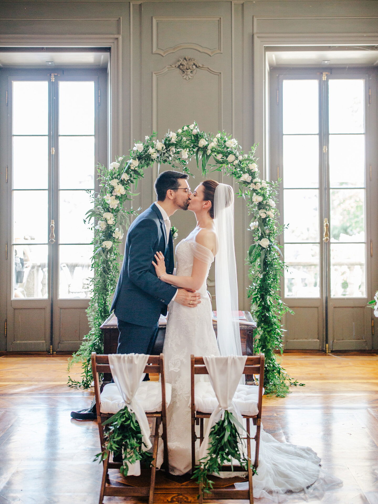Der Hochzeitskuss des Brautpaars unter einem mit Blumen und Grün dekorierten Bogen. Die Braut trägt ein schulterfreies Kleid und einen langen Schleier, während der Bräutigam in einem dunklen Anzug sie zärtlich umarmt. Große Fenster rahmen die Zeremonie ein.