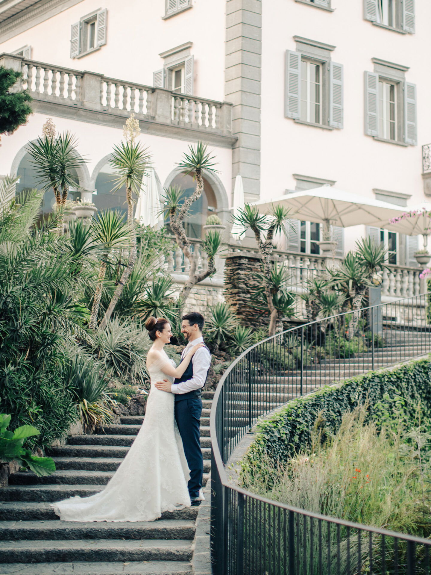 Das frisch verheiratete Paar steht auf einer von grünen Pflanzen umgebenen Treppe, die zu einer großen Villa führt. Die Braut trägt ein weißes Kleid mit langer Schleppe, der Bräutigam einen blauen Anzug. Beide blicken sich liebevoll an.