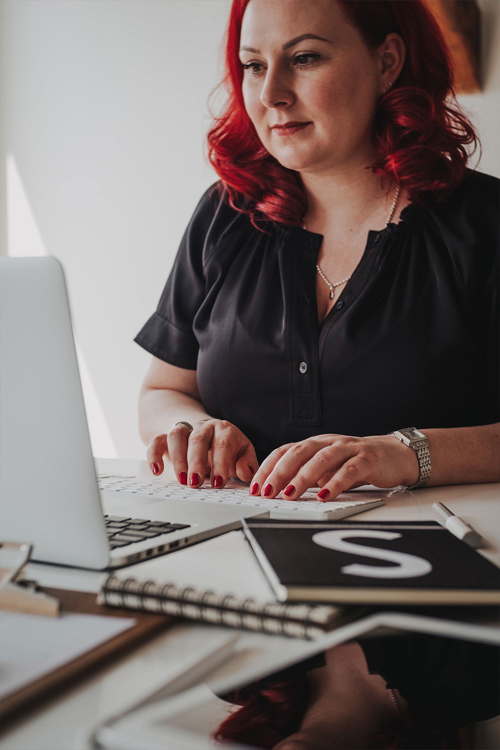 Eine Frau mit roten Locken arbeitet an ihrem Laptop und trägt eine elegante schwarze Bluse. Auf dem Schreibtisch vor ihr sind Notizblöcke verstreut.