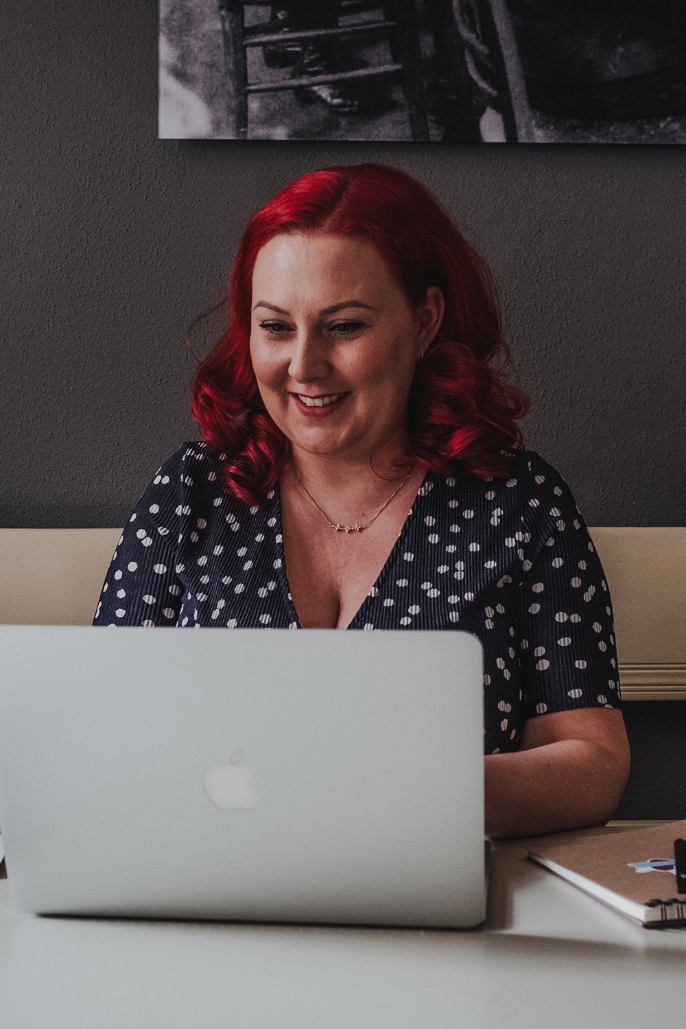 Die Frau mit roten Haaren sitzt entspannt vor ihrem Laptop und trägt ein gepunktetes Kleid. Sie lächelt zufrieden in ihrem Büro.