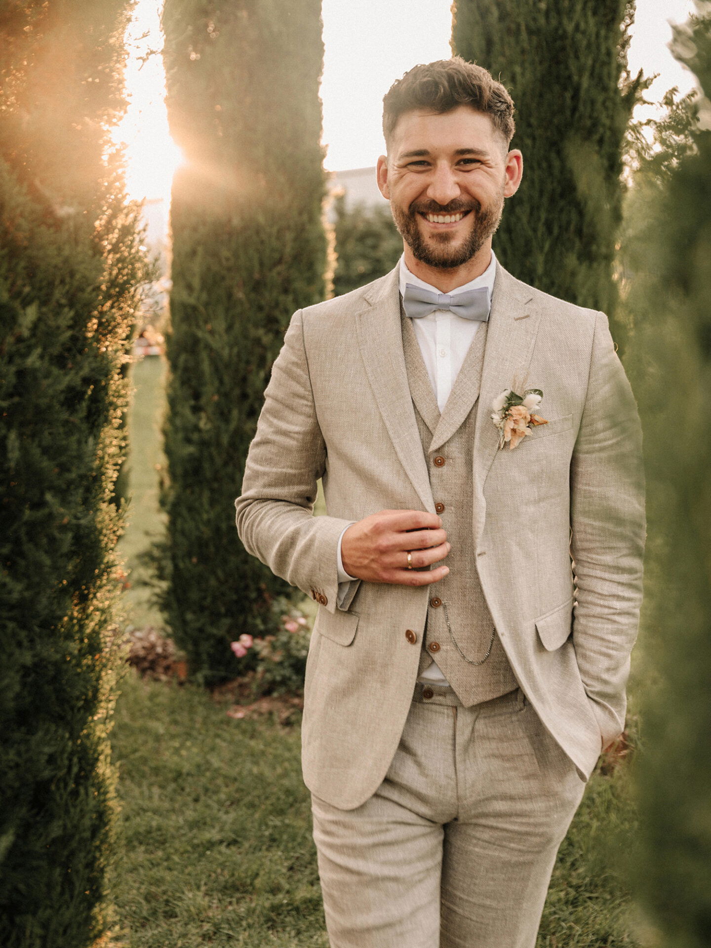 Der Bräutigam lächelt fröhlich in die Kamera, während er in einem gepflegten, hellen Anzug mit einer Fliege posiert. Die Sonne scheint durch die Bäume im Hintergrund und erzeugt eine warme, romantische Atmosphäre.