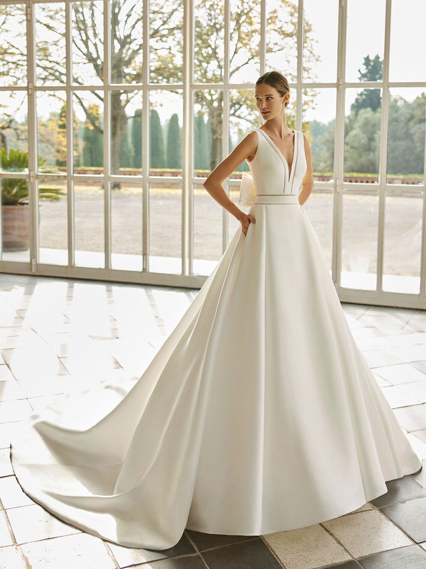Eine Frau trägt ein schlichtes, ärmelloses A-Linien-Brautkleid mit einem tiefen V-Ausschnitt. Sie steht in einem großen Raum mit Fenstern, die einen malerischen Blick auf einen Garten bieten. Der Rock ihres Kleides fließt elegant hinter ihr.