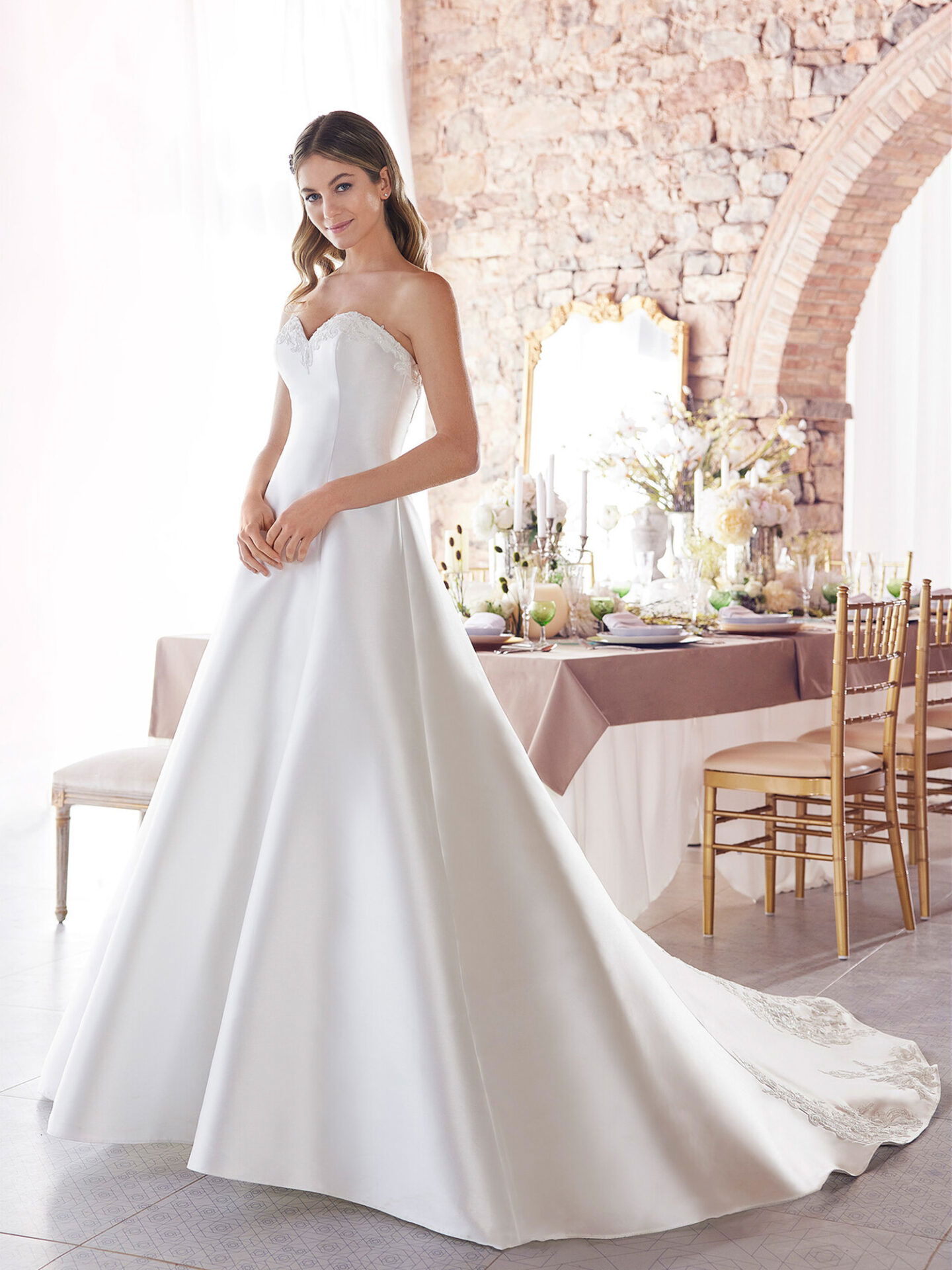 Eine Frau trägt ein trägerloses A-Linien-Brautkleid mit einem langen, fließenden Rock. Sie steht vor einem festlich dekorierten Tisch in einem stilvollen Raum mit Steinwänden und einem großen Fenster, das weiches Licht hereinlässt.