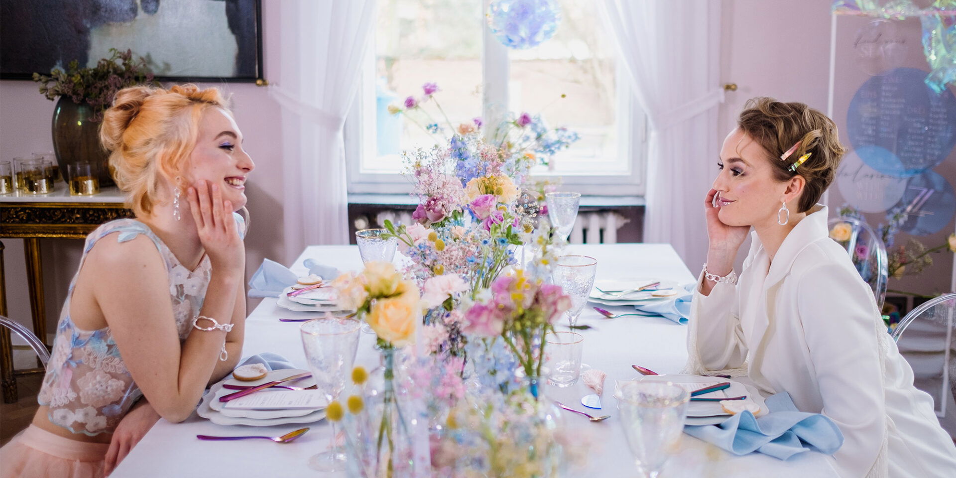Zwei Frauen an einem weiß gedeckten Tisch voller pastellfarbener Blumengestecke tauschen lächelnd Blicke aus. Der Tisch ist festlich für eine Feier oder Hochzeit arrangiert, und im Hintergrund fällt weiches Tageslicht durch ein großes Fenster, das den Raum freundlich erhellt.
