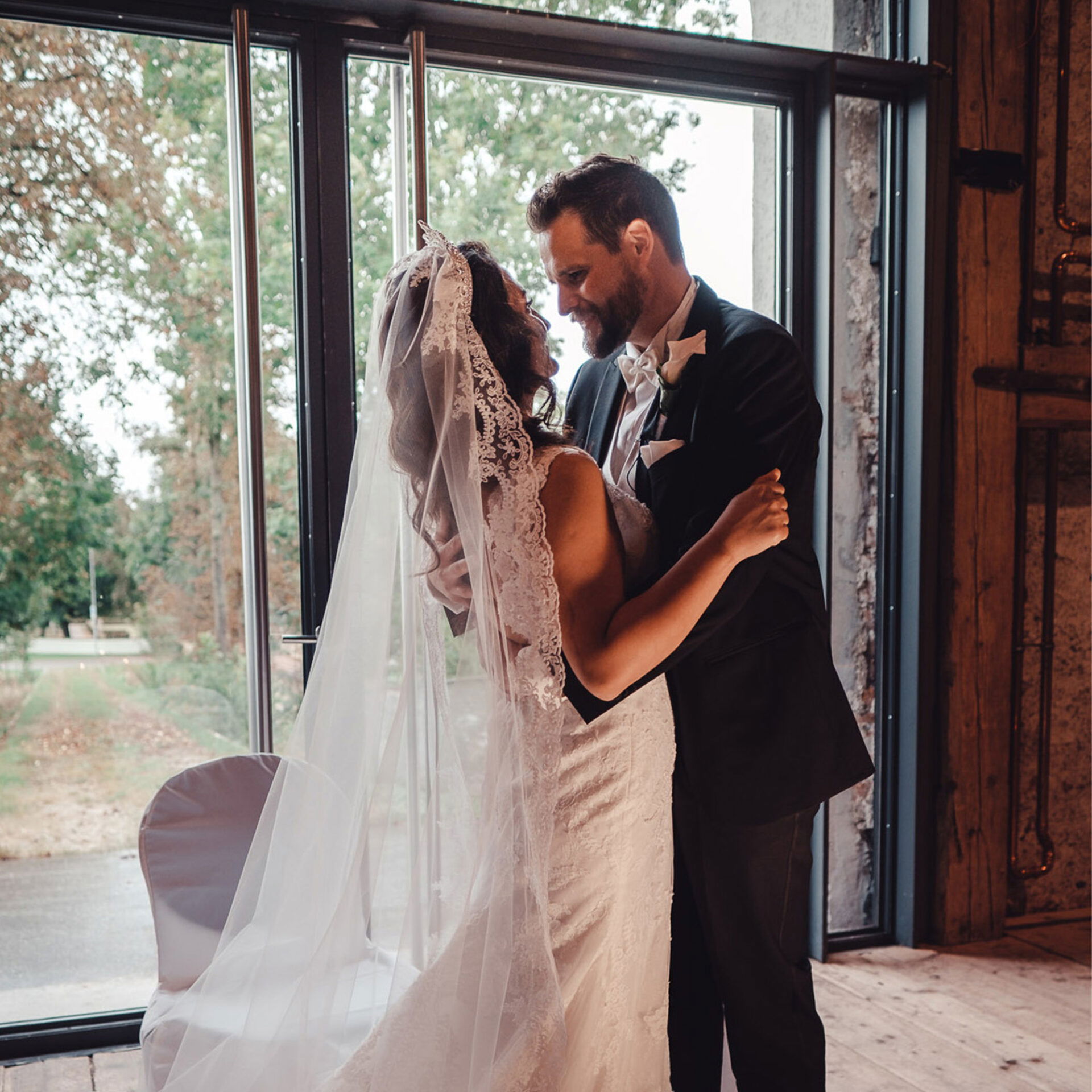 Ein emotionaler Augenblick zwischen einem frisch verheirateten Paar in einem stilvollen, modernen Raum mit großen Glasfenstern. Die Braut in einem weißen Kleid und Schleier und der Bräutigam im schwarzen Anzug stehen eng beieinander und blicken sich liebevoll an.