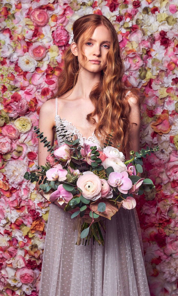 Eine Braut mit offenen, roten Locken posiert vor einer eindrucksvollen Blumenwand aus Ranunkeln in verschiedenen Rosatönen. Sie hält einen großen Blumenstrauß, der aus Ranunkeln, Orchideen und Eukalyptuszweigen besteht. Ihr zartes Kleid und die romantische Kulisse betonen den verträumten Hochzeitslook.