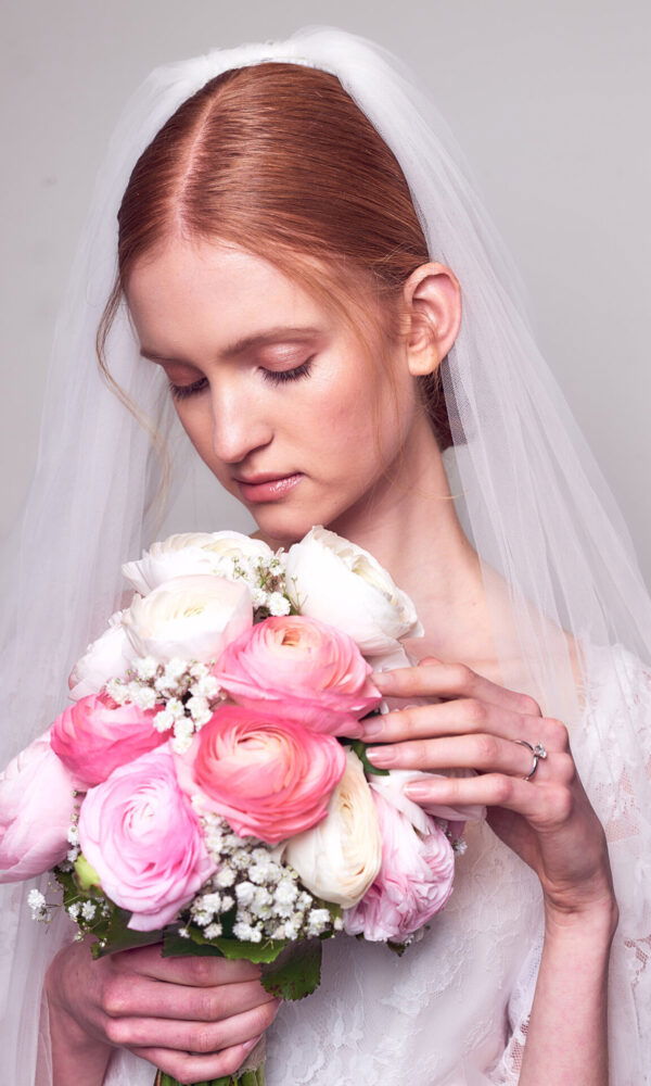 Eine Braut mit langem, glattem Haar trägt einen weißen Schleier und hält einen großen Blumenstrauß aus rosa und weißen Ranunkeln. Sie blickt zärtlich auf die Blumen in ihren Händen, während ihr schlichtes Brautkleid und der elegante Strauß die romantische Stimmung des Bildes unterstreichen.