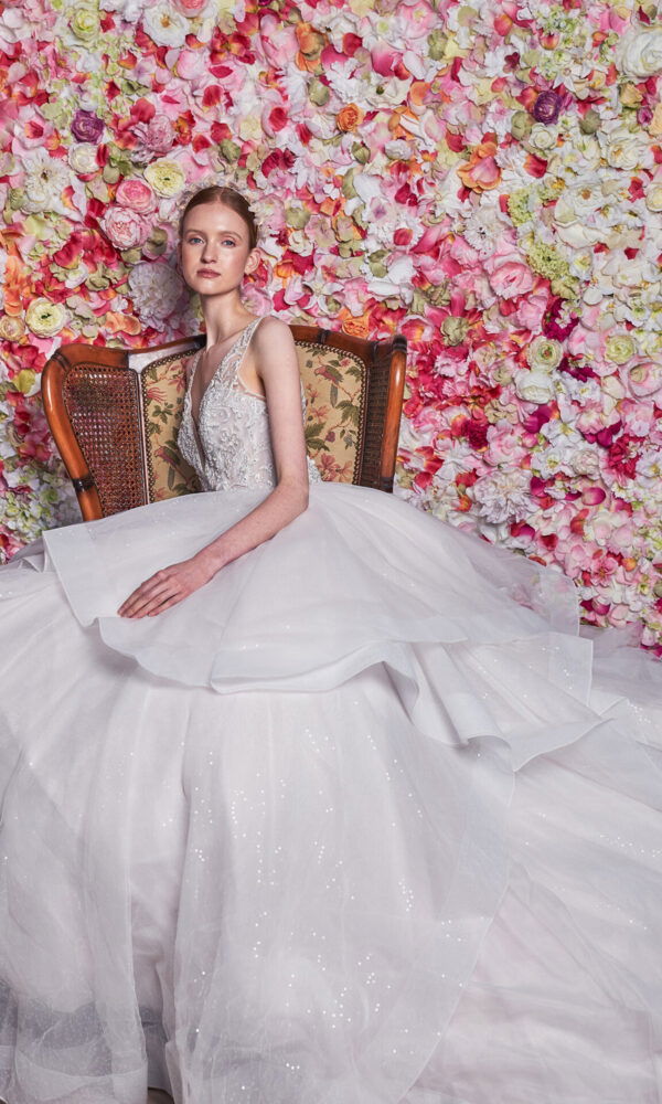 Eine Braut in einem traumhaften, weiten Tüllkleid sitzt auf einem antiken Stuhl vor einer üppigen Blumenwand aus Ranunkeln. Die Blumen in Rosa- und Cremetönen verstärken den dramatischen Look des voluminösen Kleides, das sich in weiten Schichten um sie herum ausbreitet.