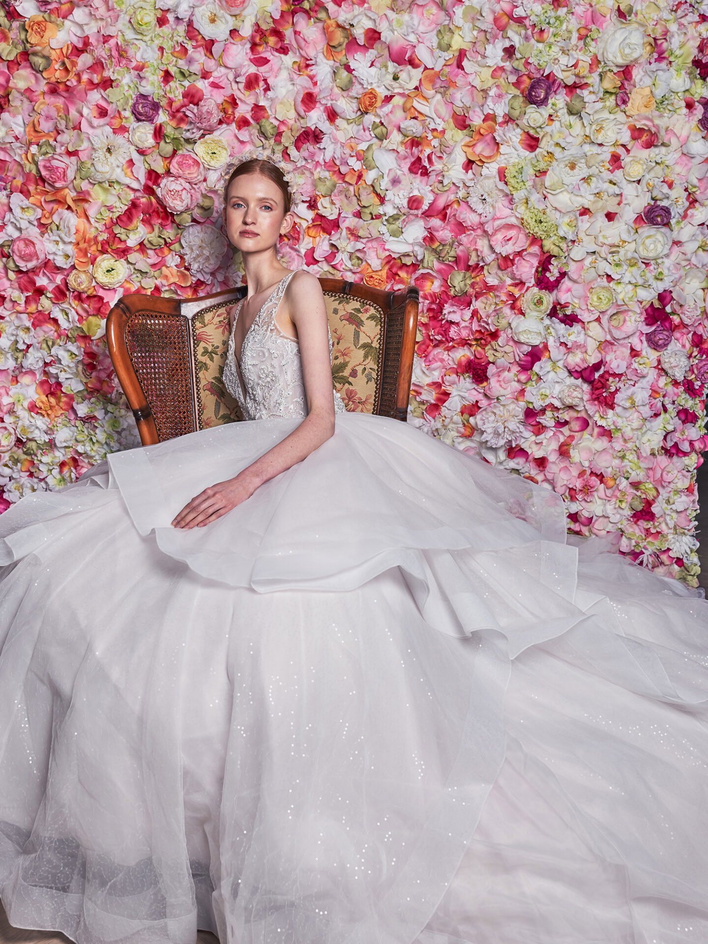 Eine Braut in einem traumhaften, weiten Tüllkleid sitzt auf einem antiken Stuhl vor einer üppigen Blumenwand aus Ranunkeln. Die Blumen in Rosa- und Cremetönen verstärken den dramatischen Look des voluminösen Kleides, das sich in weiten Schichten um sie herum ausbreitet.