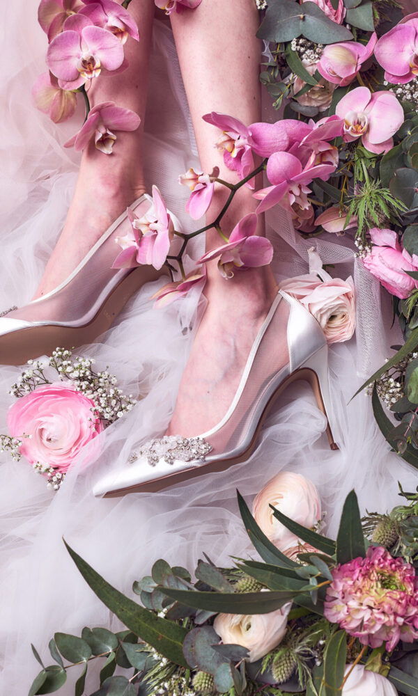 Zarte Brautschuhe auf weißem Tüll, umringt von rosa Orchideen und grünen Blättern. Die Szene wird durch Ranunkeln und andere Blumen in Pastelltönen ergänzt, die sich sanft um die Beine der Braut legen und den eleganten Stil unterstreichen.