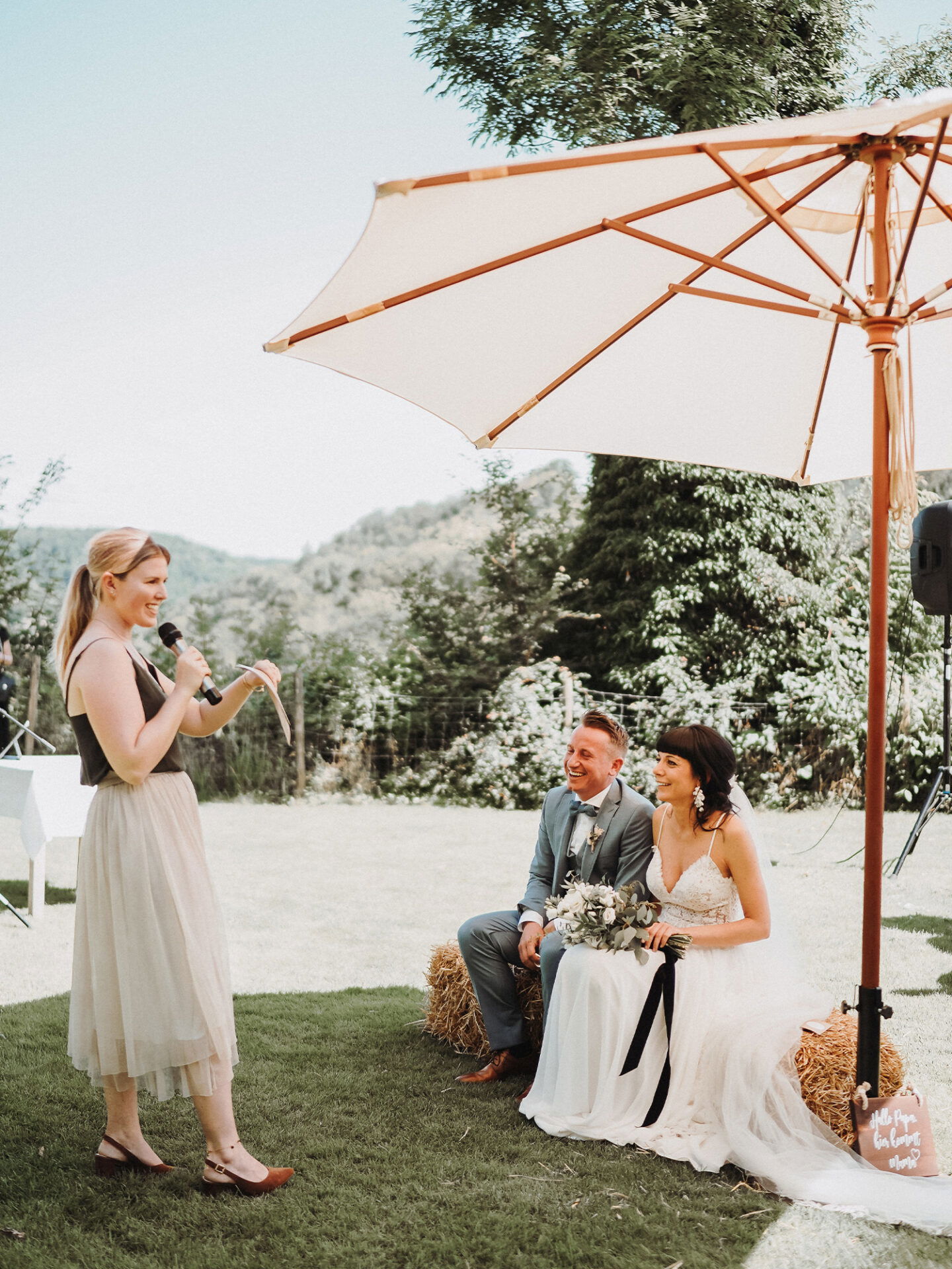 Das Brautpaar sitzt bei ihrer Outdoor-Hochzeit unter einem Sonnenschirm und hört einer Rednerin zu. Die Braut in einem weißen Hochzeitskleid und der Bräutigam in einem hellblauen Anzug lächeln. Im Hintergrund erstreckt sich eine grüne Landschaft.